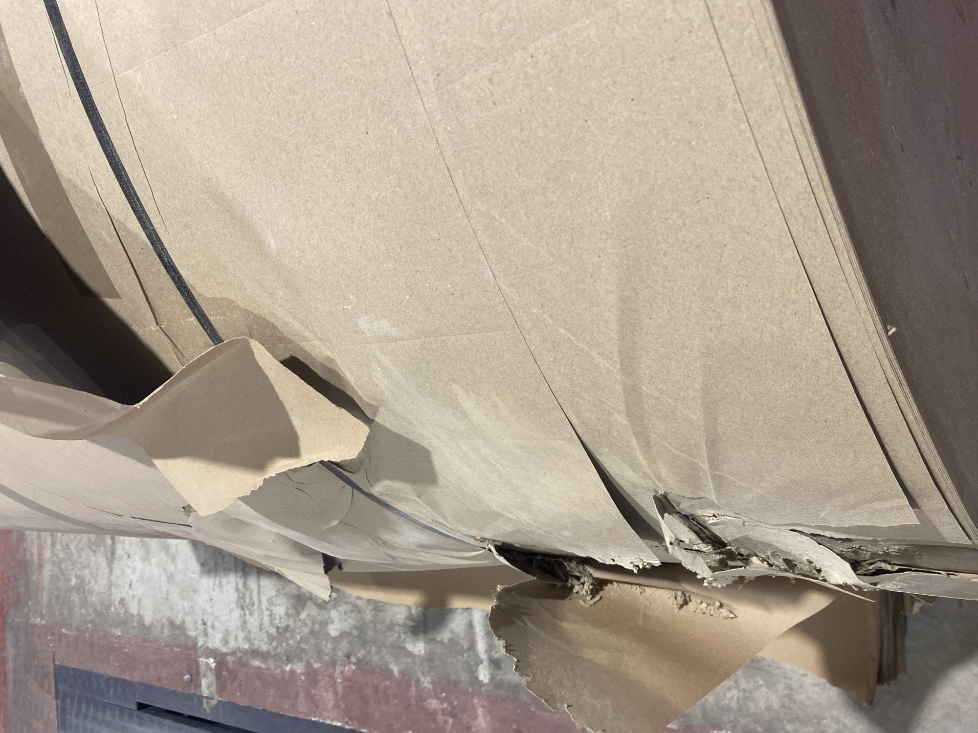 A stack of brown cardboard sheets is situated on a floor, possibly in Paipe, Nigeria, though the location cannot be definitively confirmed from the image alone. The cardboard sheets are significantly damaged, with torn and crumpled edges. A black strap runs horizontally across the sheets. The floor has a gray and red marbled pattern. A black metal structure is visible in the lower left corner of the image. There are no people visible. The environment appears to be an industrial or storage setting.