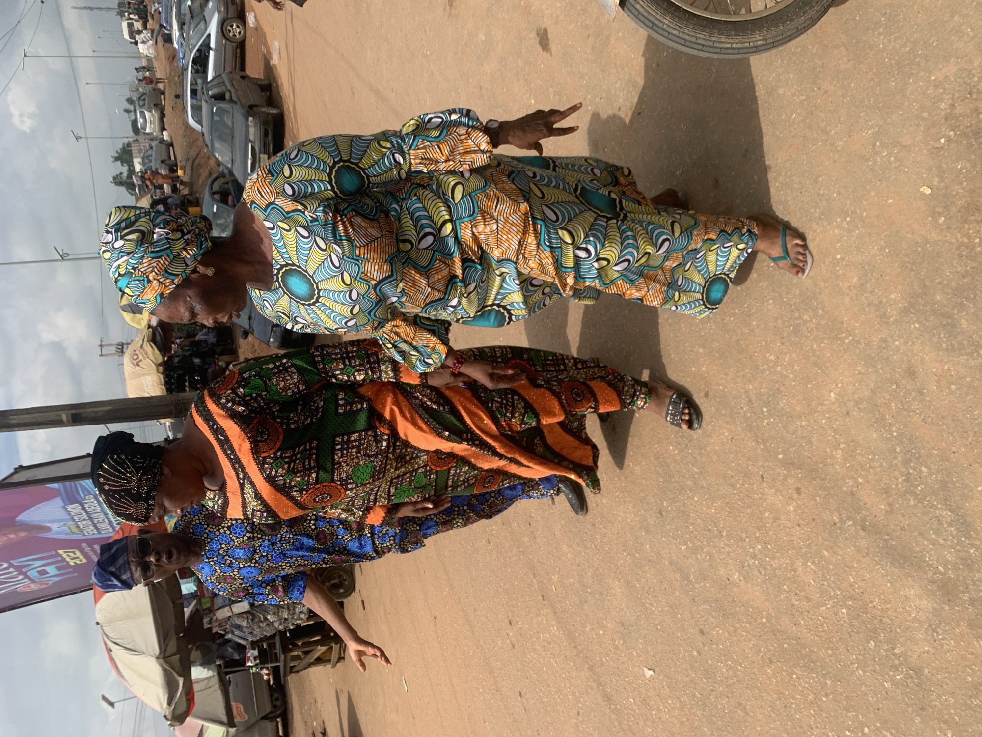 In Shagamu, Nigeria, three individuals are walking on a dirt road. A man in a blue patterned shirt and blue headscarf is on the left, gesturing with his right hand. To his right, a woman in a black headscarf and orange and green patterned dress is walking. Further to the right, a woman in a colorful headscarf and a long, patterned dress is walking, also gesturing with her right hand. In the background, there are stalls, vehicles including cars and a motorcycle, and a large billboard. The sky is cloudy.