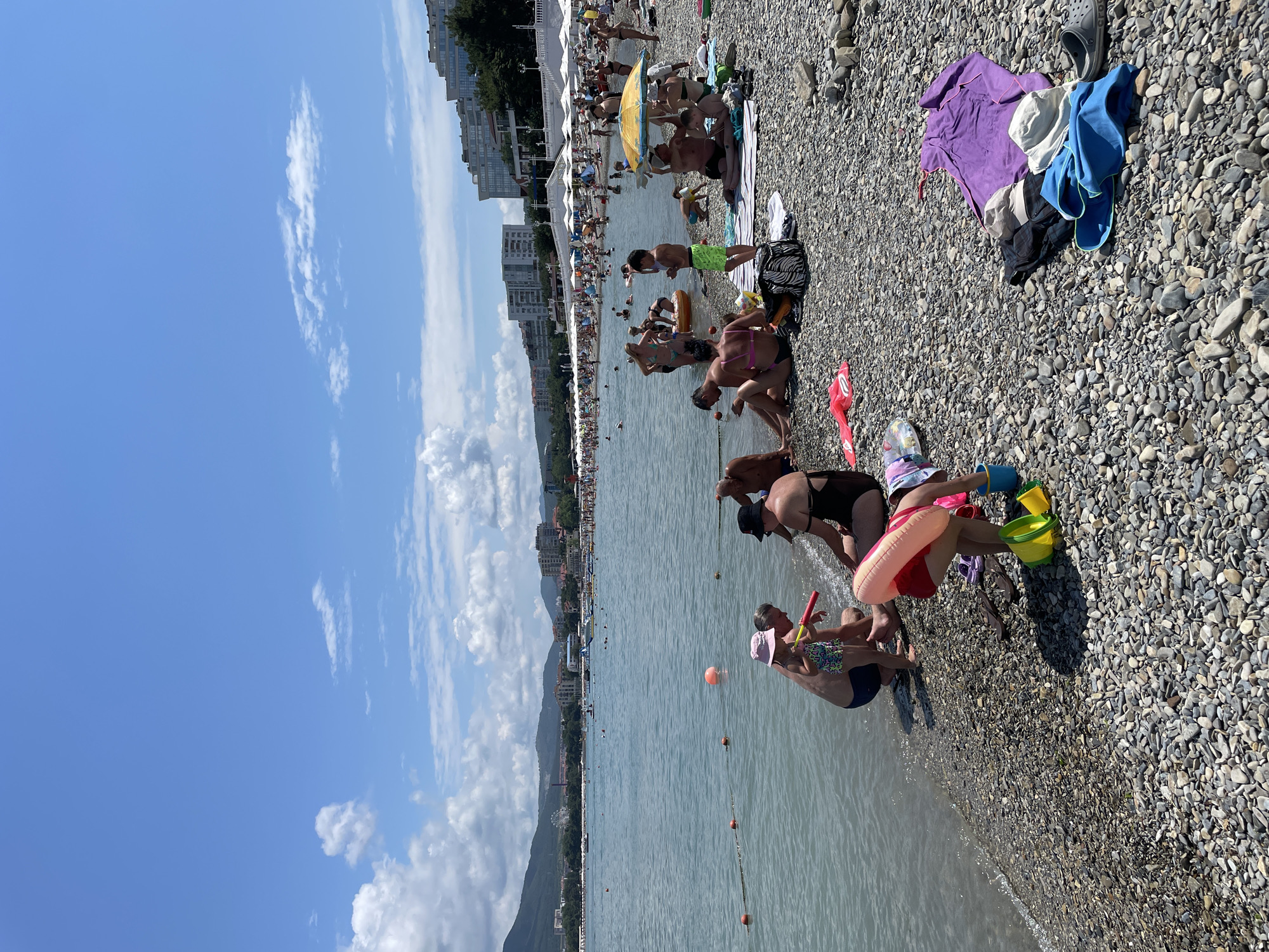 The image depicts a crowded pebble beach in Gelendzhik, Russia, on a sunny day with scattered clouds. The sea is calm and a light blue, with a rope barrier marking off an area in the water. A diverse group of people, including adults and children, are engaged in various beach activities. Some are sitting on towels and blankets, others are wading in the water, and children are playing with toys. Beach umbrellas and a few buildings are visible in the background, along with distant hills. The overall scene is one of a bustling summer day at the seaside.