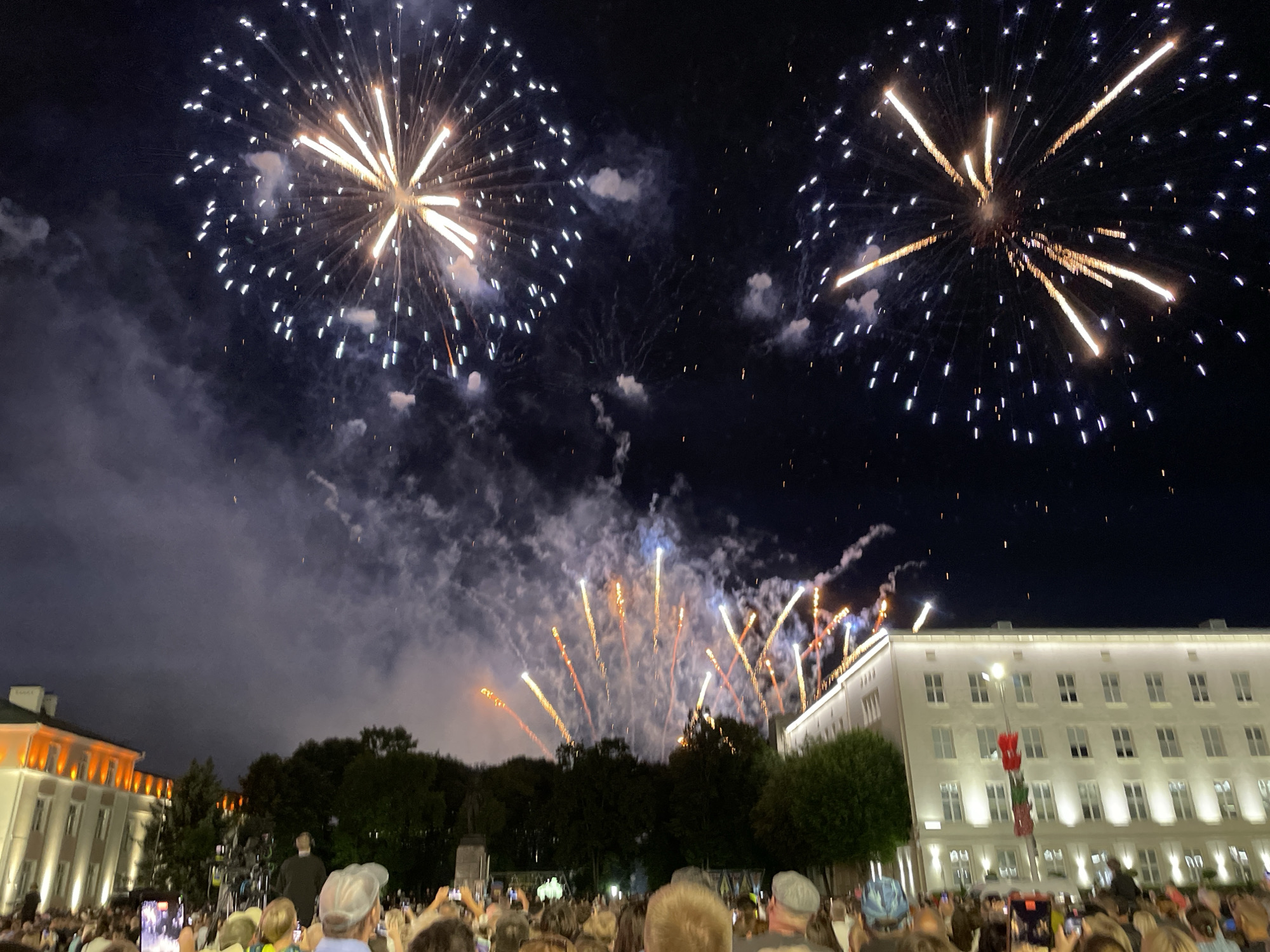 The image depicts a nighttime fireworks display over the city of Brest, Belarus. Two large fireworks are visible in the dark sky, one on the left and one on the right, with trails of light and smoke emanating from them. Below the fireworks, a crowd of people is gathered, many holding up their phones to capture the event. In the background, there are buildings illuminated with warm light, suggesting they are part of the city. Trees can be seen between the crowd and the buildings. The overall scene is festive and celebratory, with the fireworks illuminating the night sky.
