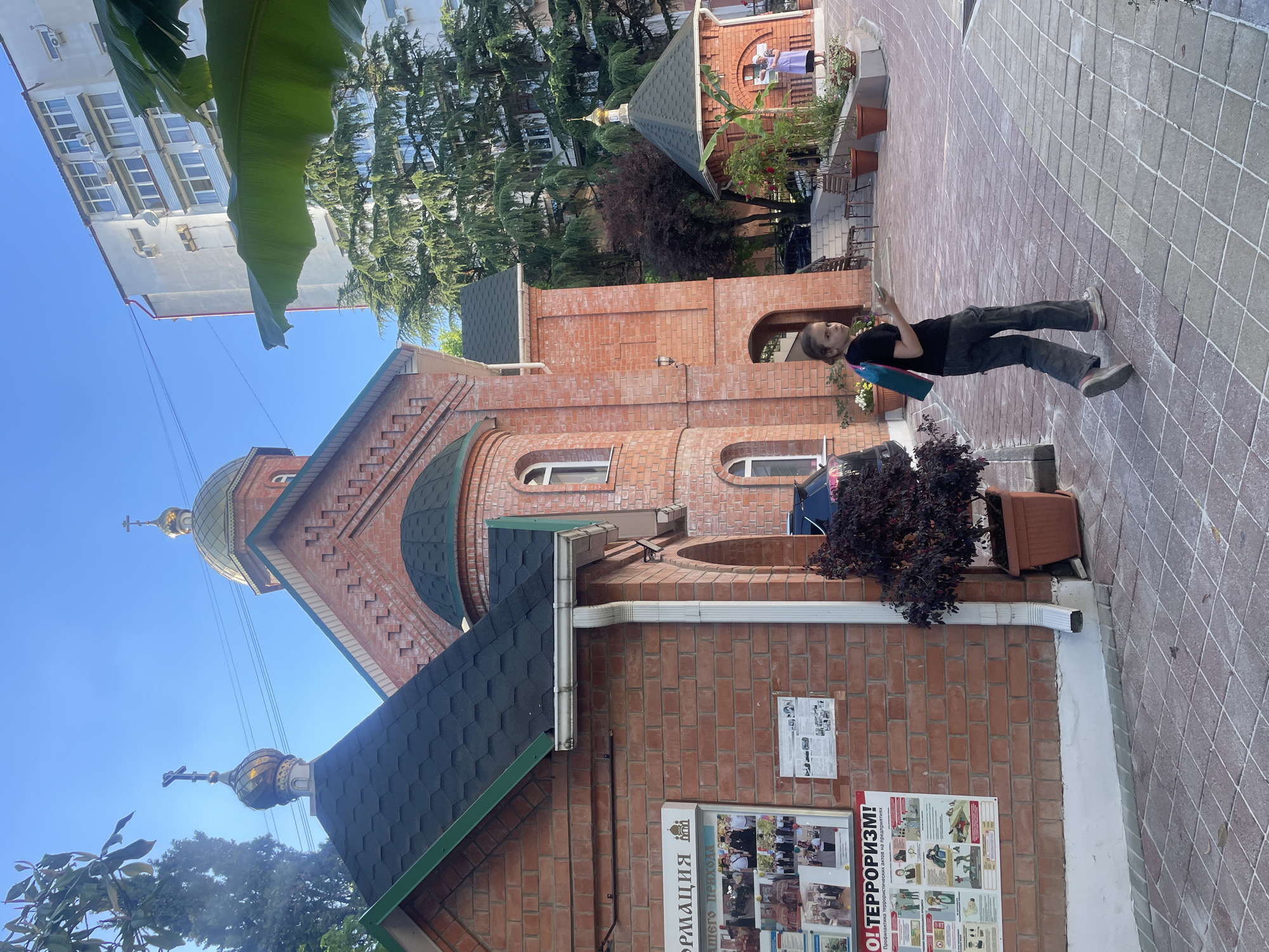 This daytime outdoor scene depicts an Orthodox church complex in Sochi, Russia. The central structure is a red brick building with a dark green shingle roof and a prominent golden onion dome topped with an Orthodox cross. A smaller, similar golden dome and cross are visible on an adjacent section of the church's roof. White gutters and downspouts are visible on the brick facade.
On the left side of the main church building, a white information board is affixed to the brick wall. Partial Russian text "ФОРМАЦИЯ" (Information) is visible at the top, and "ТЕРРОРИЗМ!" (Terrorism!) is clearly displayed in red capital letters at the bottom, indicating anti-terrorism awareness material. Various images and additional text are present on this board. A dark-leafed potted plant sits beneath the board.
In the mid-ground to the right, a smaller, brick structure with a dark green conical roof and a small golden onion dome with a cross stands surrounded by potted plants. Further back, a woman wearing a light-colored dress or apron and a dark head covering is standing near this smaller structure.
In the immediate foreground, a young person with dark hair, wearing a black t-shirt, dark pants, and light-colored sneakers, walks away from the viewer. They carry a teal or blue bag on their back and hold a light-colored rectangular object, possibly a phone, in their right hand. The person is turned slightly to their left, looking towards the left side of the frame.
The ground is paved with light-colored, square tiles arranged in a grid pattern. The sky is clear and bright blue. In the background, large evergreen trees (possibly cedars) are visible, and beyond them, a tall, multi-story white residential building with numerous windows and several external air conditioning units can be seen. Thin power lines traverse the upper left portion of the sky. A dark-colored vehicle is partially visible behind the walking person, to the left of the scene.