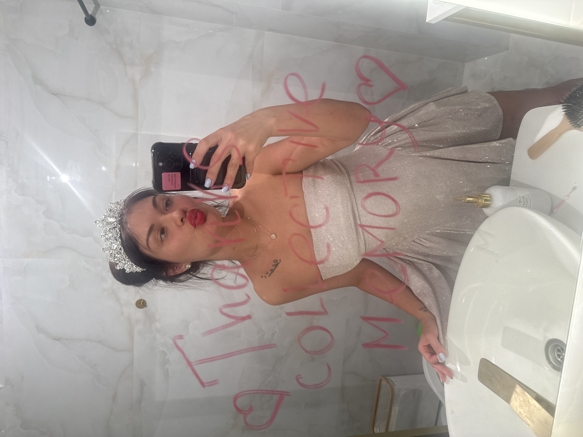 A young woman takes a mirror selfie in what appears to be a modern bathroom. The image is oriented with the woman's head to the right, as if she is looking towards the top of an upright photo. She is wearing a shimmering, strapless light-colored dress, possibly silver or champagne, and a sparkling tiara on her head, suggesting a celebratory occasion. Her dark hair is pulled back, and she has bright red lipstick, with her lips puckered. She holds a dark smartphone with a pink case in her left hand, which has light blue nail polish. A delicate gold necklace with a small pendant is visible around her neck, and there's a small tattoo on her right collarbone.
The bathroom features white and grey marble-patterned wall tiles. In the foreground, a white sink with a gold-colored faucet is visible, along with a white pump bottle (possibly soap or lotion) and a wooden-handled brush.
Written on the mirror in pink lipstick or marker, in large cursive letters, are the words "I LOVE YOU MORE" along with several heart shapes. The woman's reflection is centered in the frame, capturing her full upper body and part of her lower body. The overall mood suggests playfulness and self-adornment, likely before or after an event. The location context is Moscow, Russia.