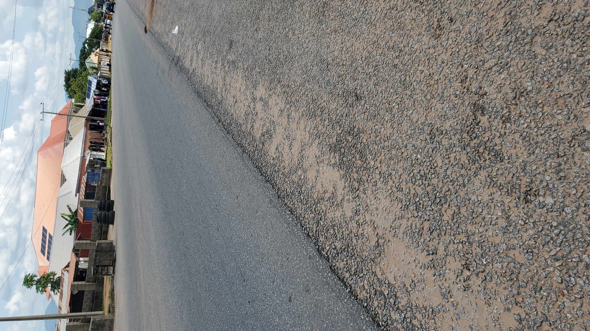 This is a medium-angle, eye-level shot of a road in Jalingo, Nigeria. The road is paved with asphalt and has a gravel shoulder. On the left side of the road, there are several buildings with red and white roofs, some with solar panels. There are also stacks of tires and what appear to be shipping containers. People are visible walking and standing in front of the buildings, some of which appear to be shops. A few vehicles, including what looks like a tricycle and a car, are on the road in the distance. The sky is cloudy with patches of blue. The overall impression is of a street scene in a town or city.