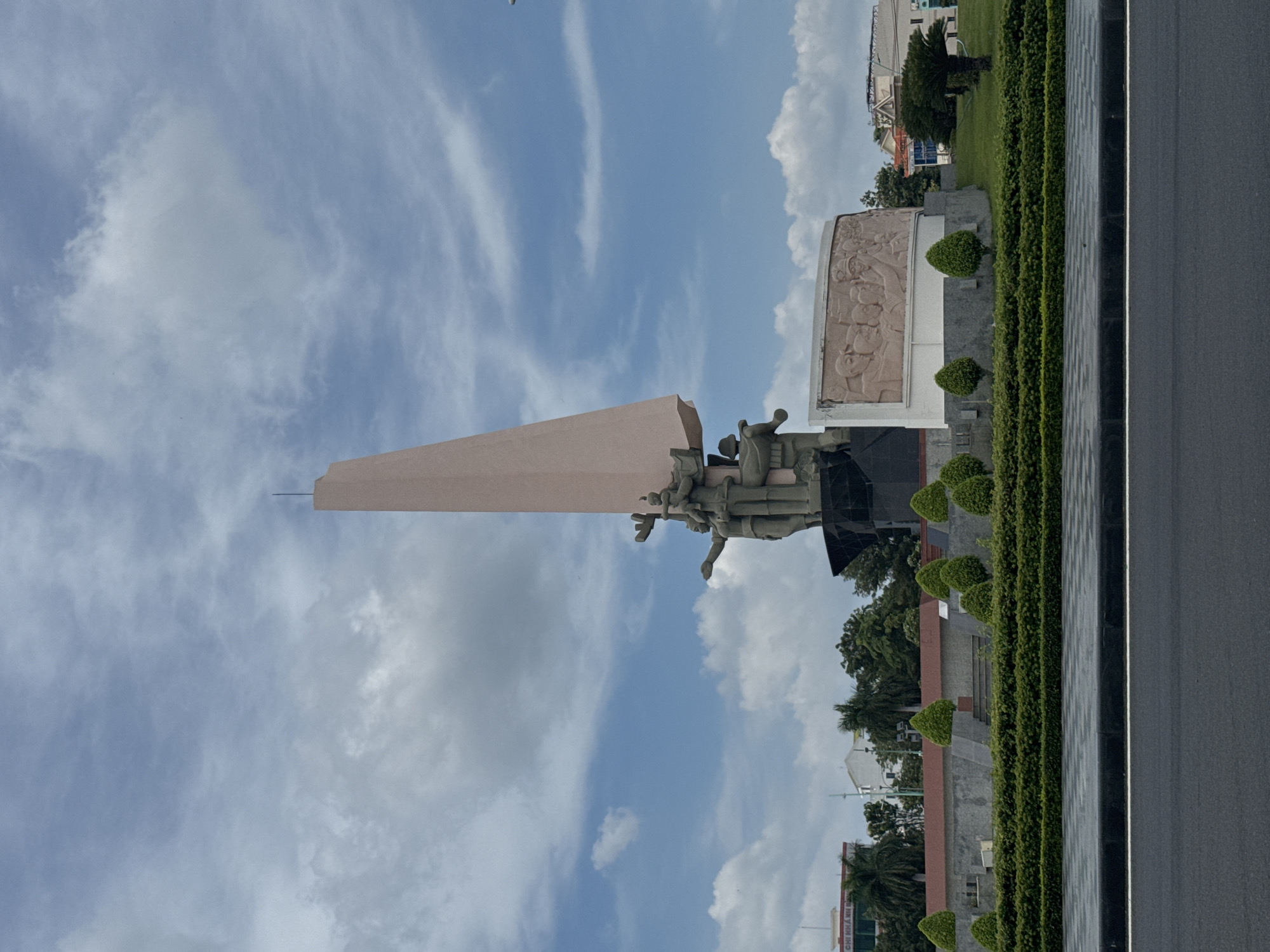 The image captures a monument located in Phan Thiết, Vietnam, under a sky with scattered white clouds against a blue background. The monument features a large, light pink obelisk-like structure at the top. Below it, a dark stone sculpture depicts several figures in dynamic poses, seemingly engaged in a collective effort. To the right of the main sculpture, a beige relief panel displays a scene with multiple figures carved in a historical or narrative style.
The monument is surrounded by meticulously maintained landscaping. A low, dense hedge forms a perimeter around the monument's base, interspersed with manicured cone-shaped trees. Beyond the hedge, a grey stone wall and a staircase are visible, leading up towards the monument. The ground in front of the monument is a paved area with a checkered pattern. A dark asphalt road with a white line runs across the foreground, indicating a street or thoroughfare.
In the background, several buildings are visible, including what appears to be a multi-story building with signage that includes Vietnamese characters. Palm trees and other lush green foliage are scattered throughout the scene, suggesting a tropical or sub-tropical environment. The overall impression is one of a public space dedicated to commemoration, set within a well-kept urban landscape.