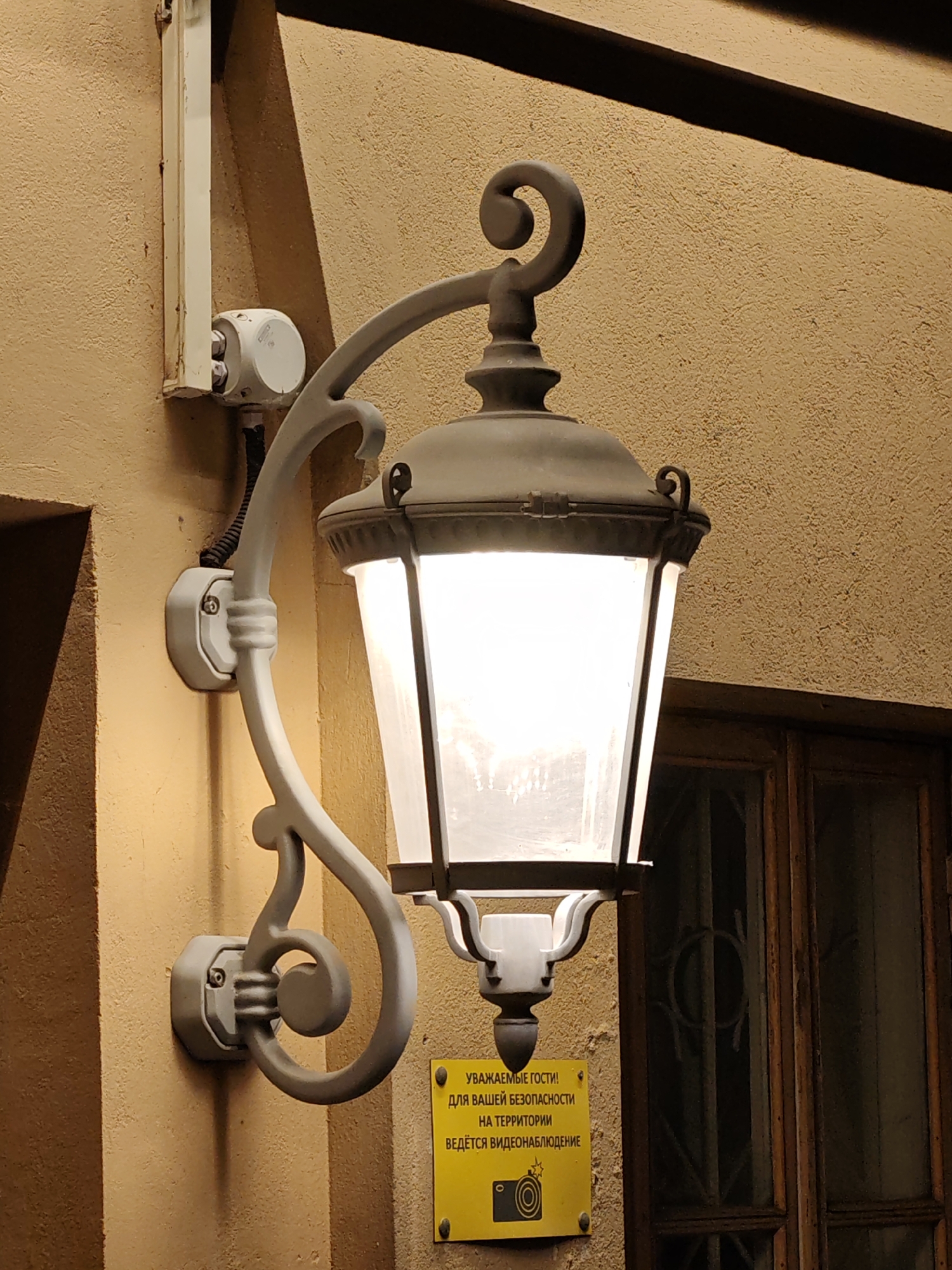 A lit, wrought-iron street lamp is mounted on the exterior of a tan-colored building, in Moscow, Russia. The lamp has a decorative arm and is attached to the wall via two mounting plates. Electrical conduit and a junction box are above the lamp. Below the lamp, on the wall, is a yellow sign in Russian warning of video surveillance in the area. To the right of the lamp is a dark-colored wooden window frame. The building facade appears textured.