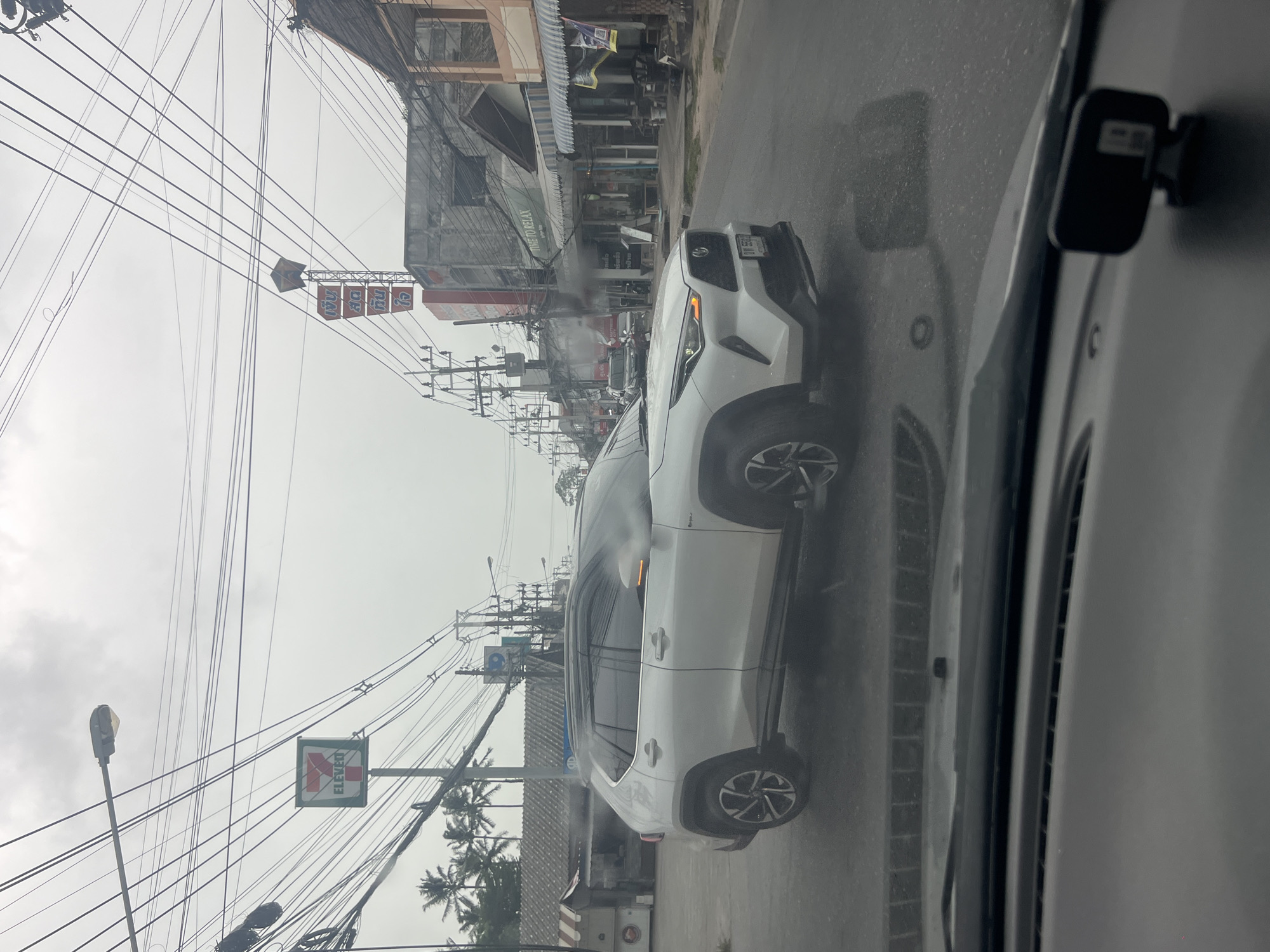 The image depicts a street scene in the city of Ko Samui District, Thailand, on an overcast day. In the foreground, the interior of a car is visible, including the dashboard and a small black device mounted on it. A white SUV is prominently featured in the middle ground, positioned on the right side of the road and appearing to be in motion. The street is wet, suggesting recent rain.
Along the sides of the road, there are various buildings, including storefronts with awnings. A "7-Eleven" convenience store sign is visible on the left, and a multi-story building with advertisements or signage in Thai script is present on the right. Overhead, a complex network of power lines and streetlights crisscrosses the sky. Palm trees can be seen in the background on the left. The overall atmosphere is somewhat hazy, possibly due to the weather or exhaust fumes.