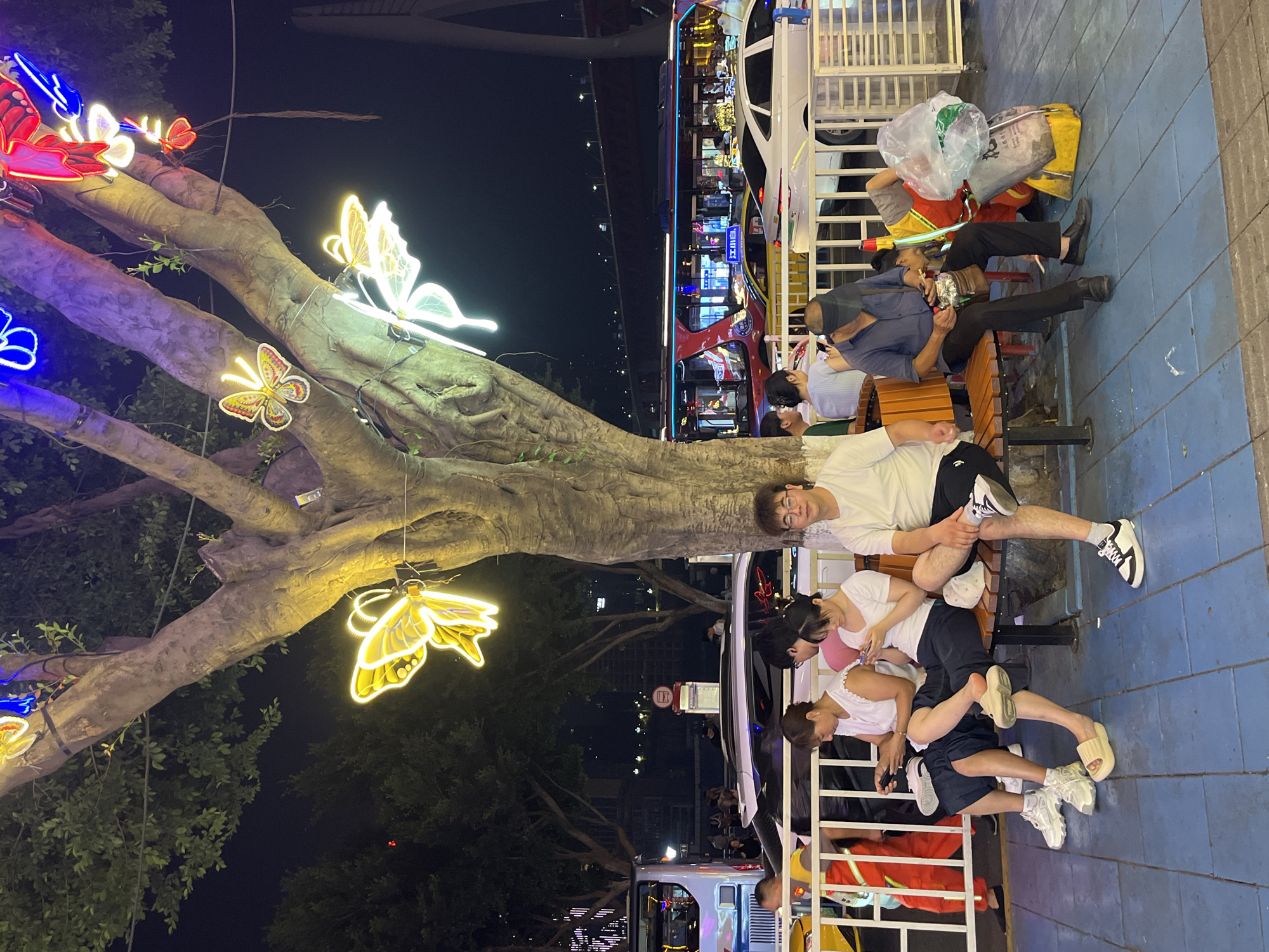 At night in Yuzhong District, China, several people are seated on a bench in front of a tree decorated with illuminated butterfly-shaped lights. The individuals include a man in a white t-shirt and black and white sneakers, a woman in a white top and dark pants, another woman in a white top and dark pants, and a man wearing a dark shirt, black pants, and a visor. A worker in a bright orange and yellow uniform is standing to the right of the bench, sorting through a pile of trash bags. In the background, various vehicles are visible, including a white car, a yellow car, and a red bus with illuminated windows. The scene is lit by the glow of the butterfly lights and the surrounding city lights.
