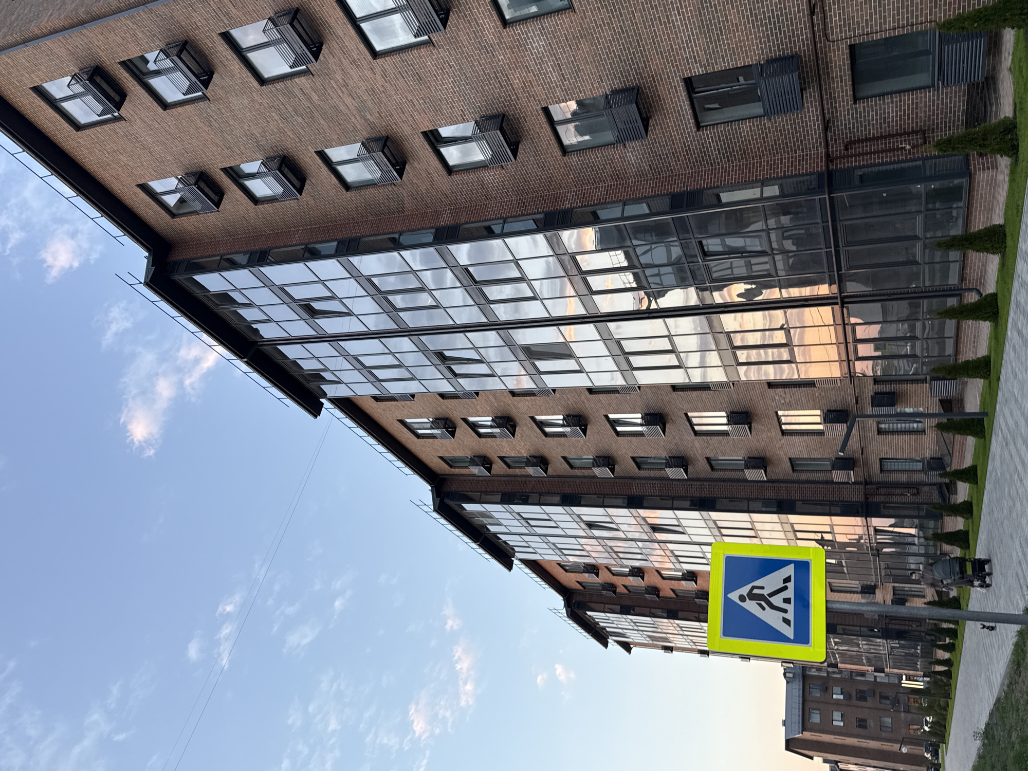 The image shows a modern brick building in Severnyi, Russia, viewed from a low angle. The building has a facade of brick with numerous windows and a glass extension reflecting the sky. A pedestrian crossing sign is prominently displayed in the foreground. The sky is blue with scattered clouds. Some greenery is visible along the edge of a paved area.