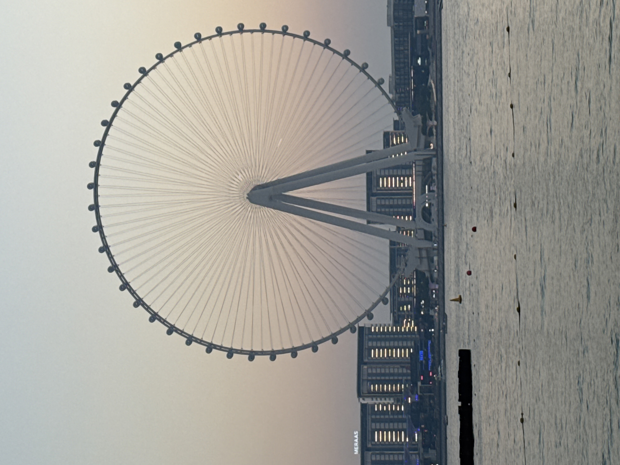 The photograph captures the Ain Dubai observation wheel, the world's largest, situated on Bluewaters Island in Dubai, United Arab Emirates. The wheel is prominently featured against a soft, dusky sky. Its massive structure, composed of a white A-frame support and a circular rim dotted with gondolas, dominates the frame. The spokes of the wheel are clearly visible, radiating from the center to the outer edge. In the background, the cityscape of Dubai unfolds, with modern high-rise buildings displaying illuminated windows. The "MERAAS" logo is visible on one of the buildings, indicating the development company. The foreground shows a body of water, likely the Persian Gulf, with several buoys and a dark, rectangular floating object, possibly part of a barrier or walkway. The scene is serene, suggesting either sunrise or sunset, with a gentle haze in the atmosphere. No people are visible in the image.