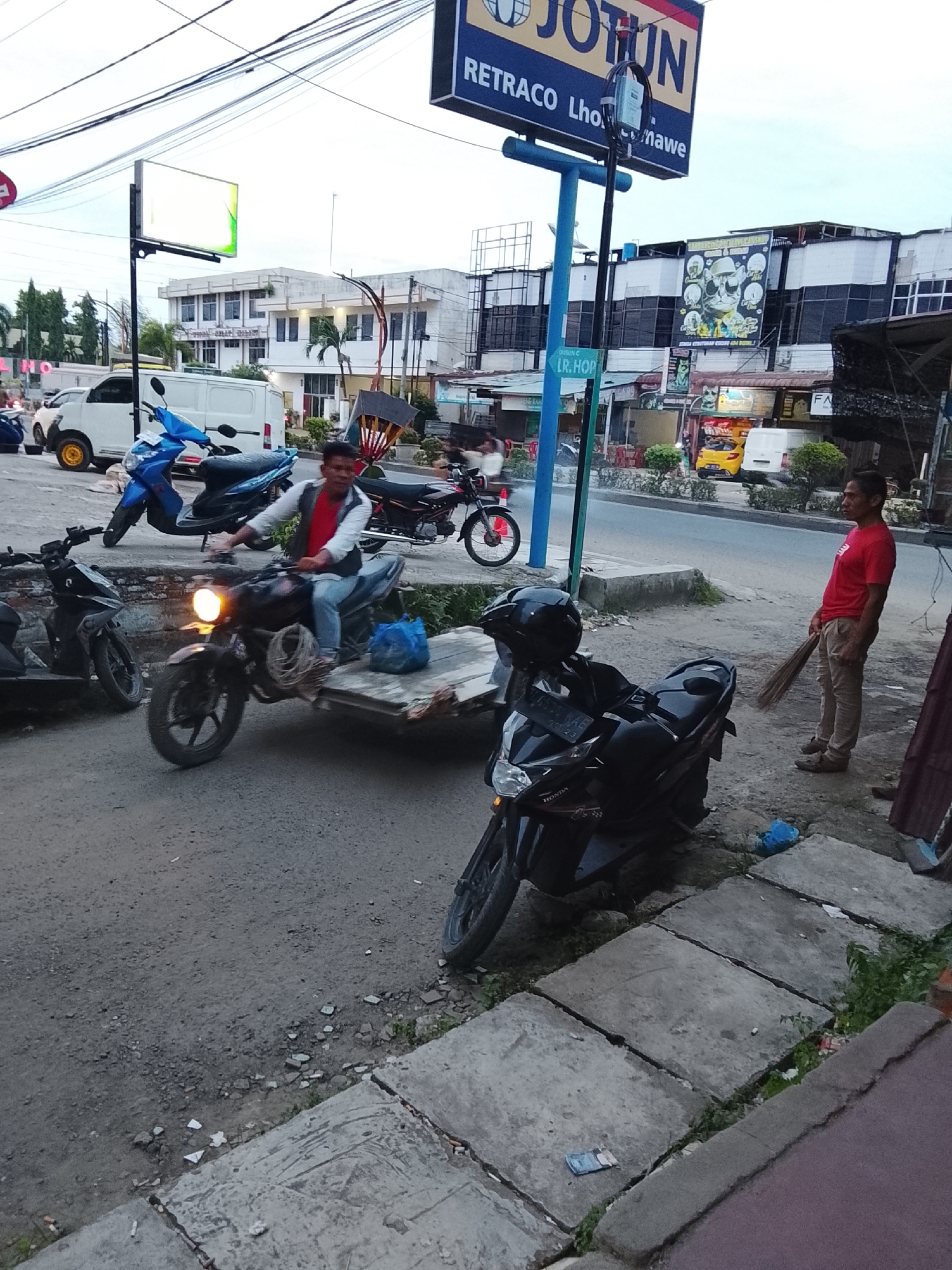 In the city of Lhokseumawe, Indonesia, on a street, a man in a red shirt and vest is riding a motorcycle. He is pulling a wooden cart filled with coiled rope. To his left, a blue scooter is parked. In the background, a white van is also parked. Further in the background, buildings with signs are visible, including a large sign for "JOTUN" and another with an image of a cat. On the right side of the image, another man in a red shirt and khaki pants is standing, holding a broom. A black motorcycle with a helmet on it is parked in front of him. The ground is a mixture of asphalt and concrete. The sky is overcast, indicating it is likely late afternoon or early evening.