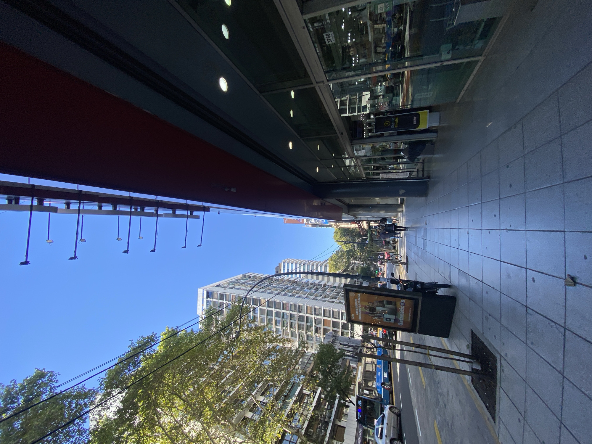 The image shows an urban street scene in Buenos Aires, Argentina. The perspective is from a low angle, looking down a city street with buildings on either side. On the right, a modern building with a glass facade and internal lighting. On the left, a building with a red exterior and overhead lighting fixtures. Below, are overhead wires visible against a clear blue sky. Trees line the street and there is street-level furniture and signage. In the distance, there are cyclists, pedestrians, and vehicles visible, suggesting an active urban environment.