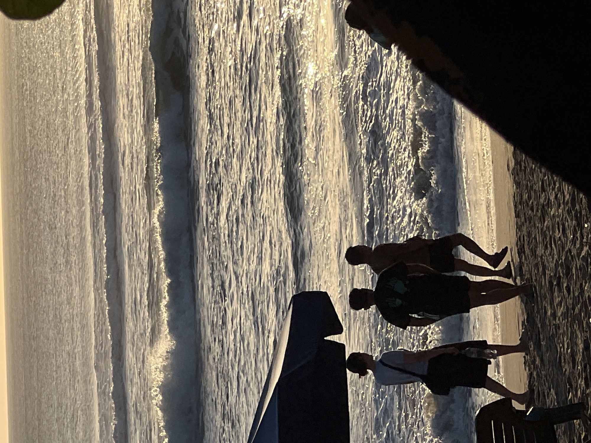 In Kuta, Indonesia, a beach scene unfolds as three individuals walk along the sandy shore, their silhouettes defined against the shimmering ocean. The sun, low in the sky, casts a warm, golden glow across the water, creating a dazzling effect as waves gently roll in. The ocean's surface is a tapestry of ripples and crests, with a few larger waves breaking near the shore. To the left, the dark canopy of a beach umbrella provides a stark contrast to the sunlit environment. The sand beneath their feet is textured, showing the footprints of those who have passed before. The overall atmosphere is tranquil and serene, capturing a moment of quiet enjoyment on a beautiful Indonesian beach.