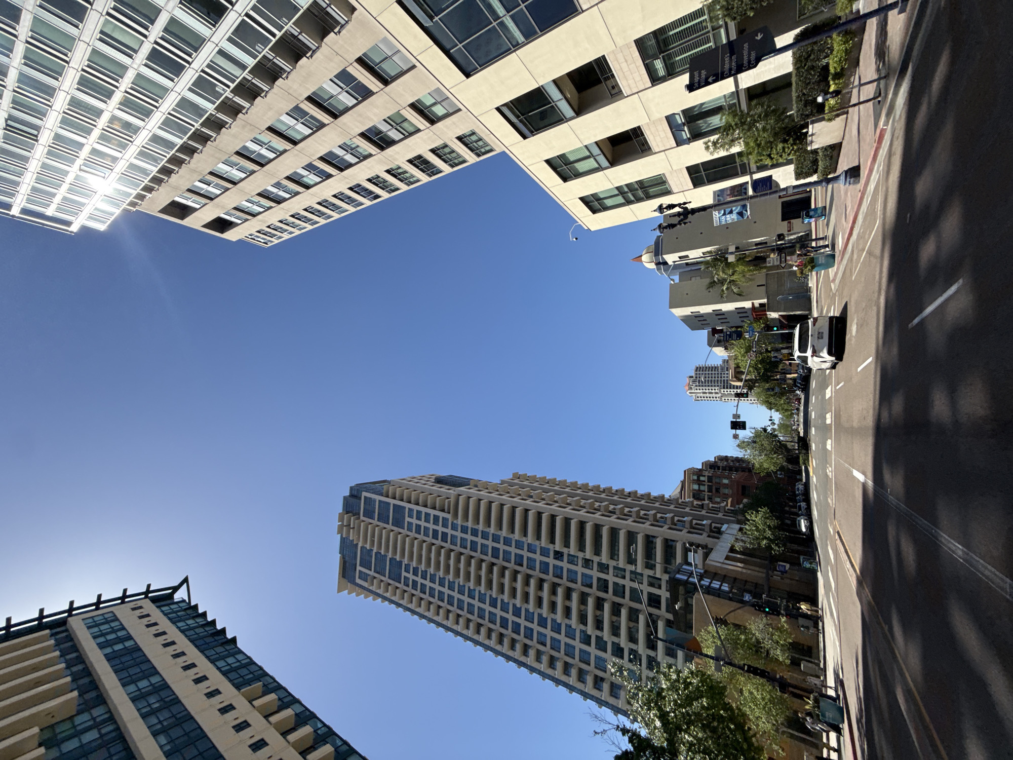 The image shows a street in San Diego, California, looking up at the surrounding buildings against a clear blue sky. Buildings of various sizes and architectural styles frame the view. On the right side of the image, there is a street with pedestrian signals and street signs, including one indicating directions to "Seaport Village" and the "Children's Museum / Convention Center." White vehicles are visible on the road. The buildings are modern high-rises, displaying repetitive window patterns and architectural details. A street lamp is also visible on the side of the road.