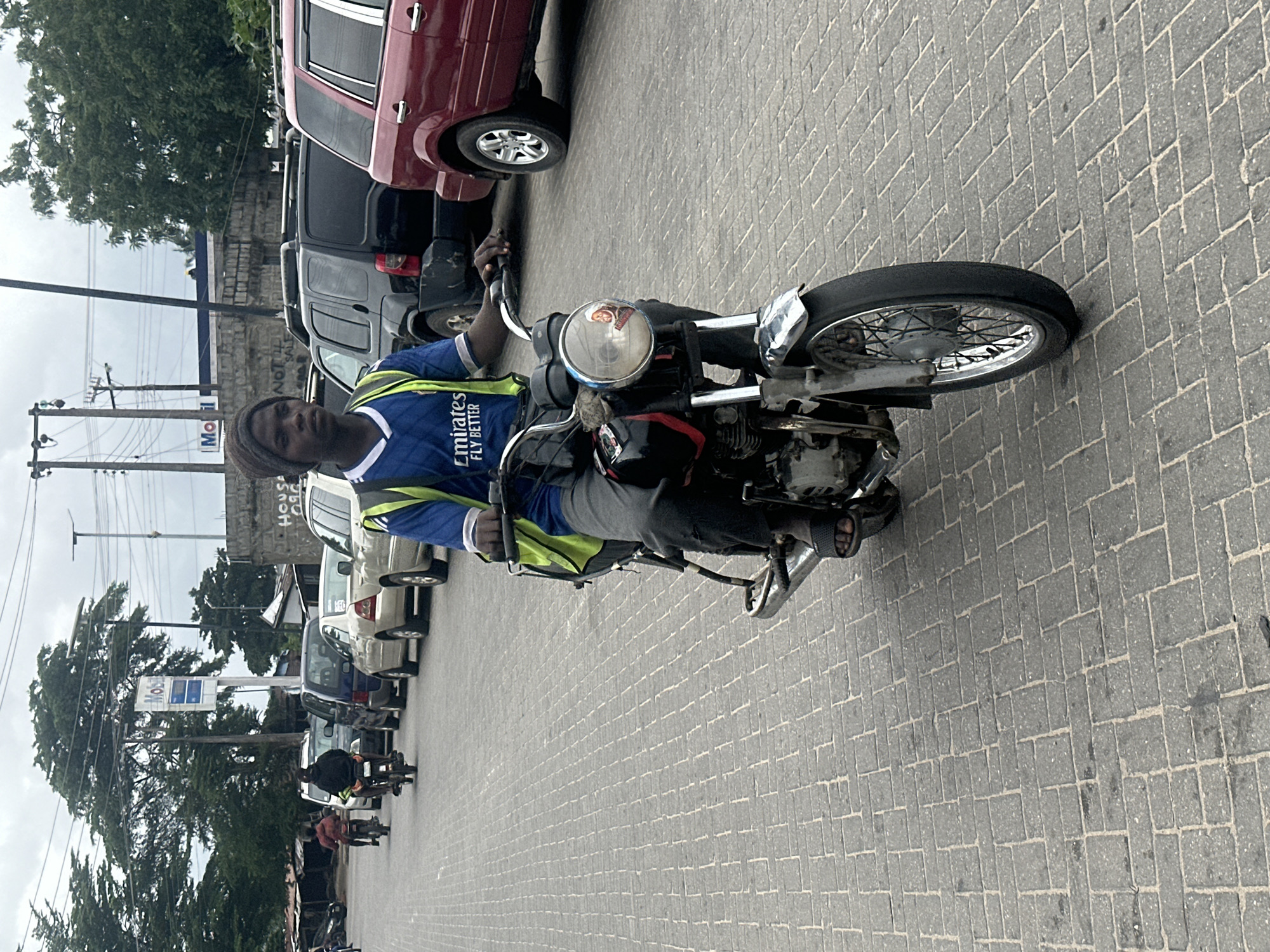 An eye-level, medium shot captures a young Black male riding a motorcycle on a paved street in Badagry, Nigeria. He is wearing a dark beanie, a blue t-shirt with white trim and "Emirates Fly Better" printed on the front, and a neon yellow vest. His legs are clad in dark pants and he wears black sandals. The motorcycle, a dark color with silver accents, features a round headlight with a circular sticker depicting a person, a neon yellow reflective vest on the handlebars, and a visible kickstand. In the background, a line of parked cars, including a red SUV and a black SUV, are visible. Beyond the cars, a Mobil gas station sign is prominent, with other buildings and lush green trees completing the urban landscape. The sky is overcast.