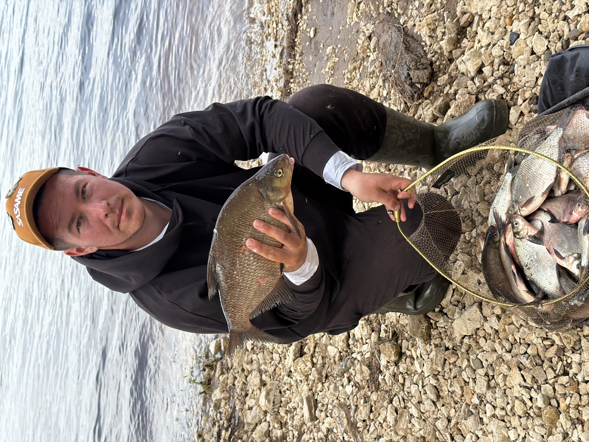 A man squats on a rocky shore, holding a fish in his hands. He wears a brown cap, a dark hooded sweatshirt, and green boots. A net full of fish rests next to him on the ground. The shore meets a body of water, which reflects the sky. This scene appears to be located near Noviy, Russia.
