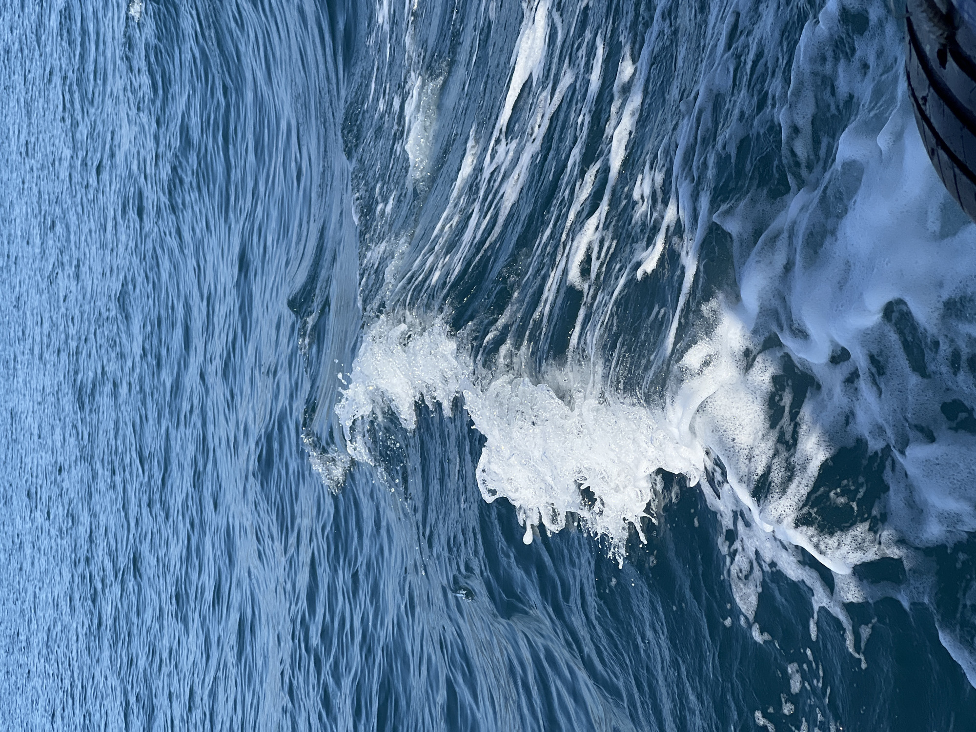 The image depicts a close-up view of the ocean surface, possibly in Nha Trang Ward, Vietnam, based on the location provided. The water is a mix of deep blue and lighter blue hues, indicating depth and surface agitation. A breaking wave is visible, characterized by white foam and splashes. The wave's texture is complex, with varying degrees of aeration and turbulent flow. A dark object, possibly a tire or part of a vessel, is visible in the upper right corner of the image. The background displays water patterns with streaks and ripples, suggesting wind or current action. There are no people visible in the image.