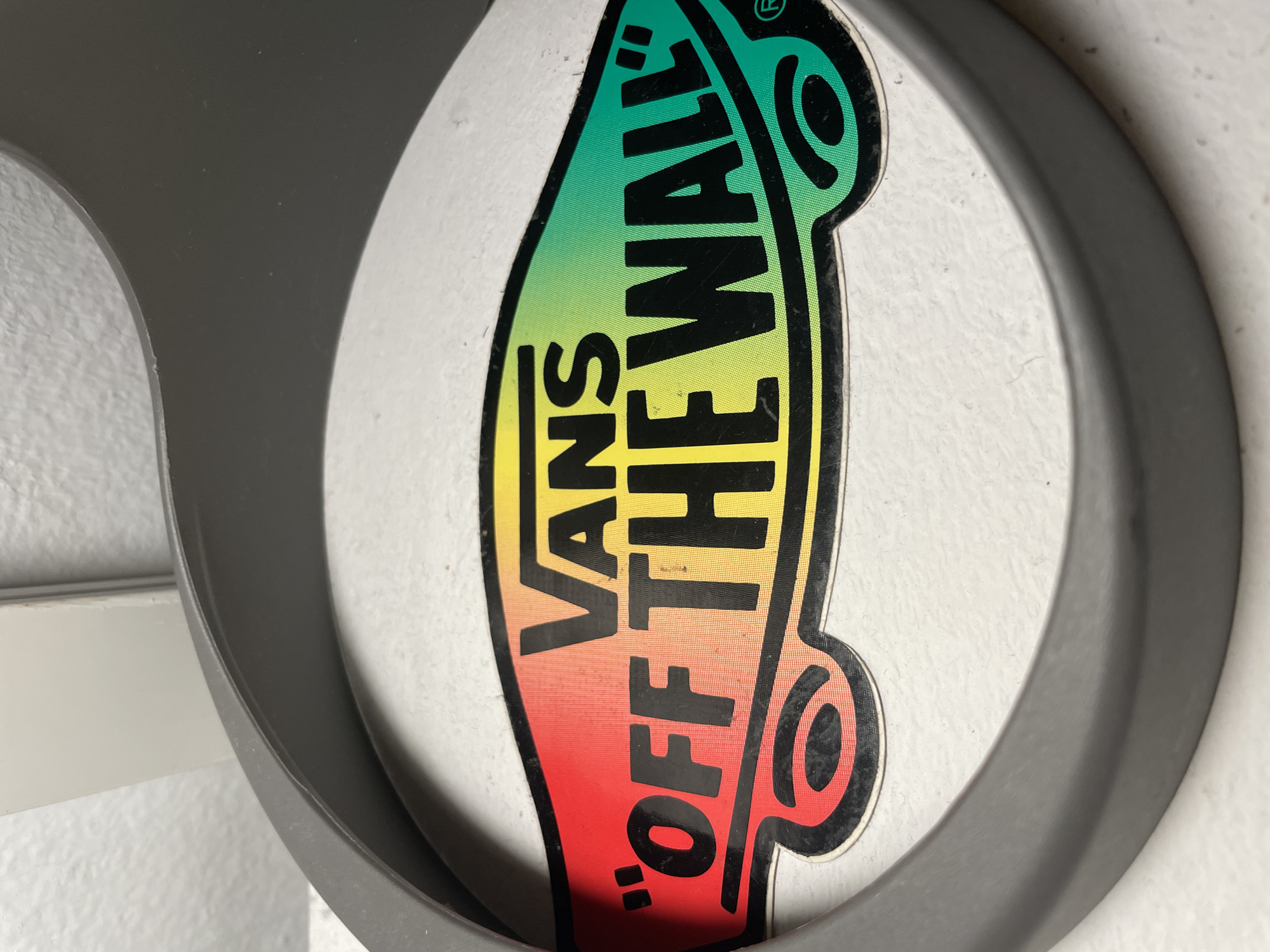A close-up shot reveals a circular Vans "Off The Wall" sticker. The sticker features a gradient color scheme, transitioning from red at the bottom to yellow and then green towards the top. The "Vans" and "Off The Wall" text are printed in black. The sticker is adhered to a white surface and is partially framed by a gray, plastic, circular object. The specific location in Barcelona, Spain is not discernible from the image.