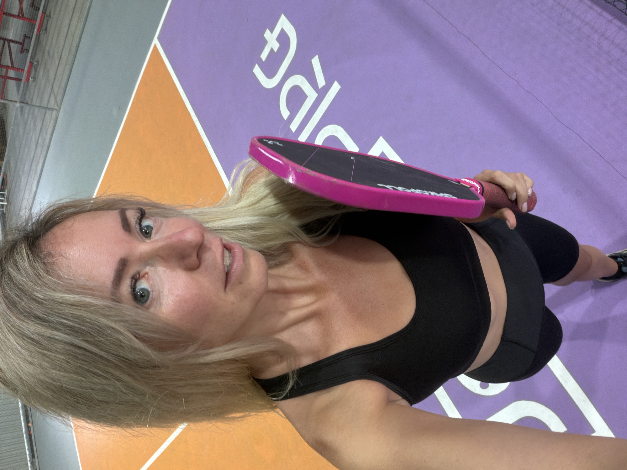 A blonde woman in athletic wear holds a pink paddle over a purple court with white markings. The woman is looking towards the camera, and her right hand is visible holding the paddle. The court is marked with the word "DALAT" in large white letters. The background shows an orange court area and a gray industrial-looking ceiling with red equipment. The scene appears to be indoors at a sports facility.