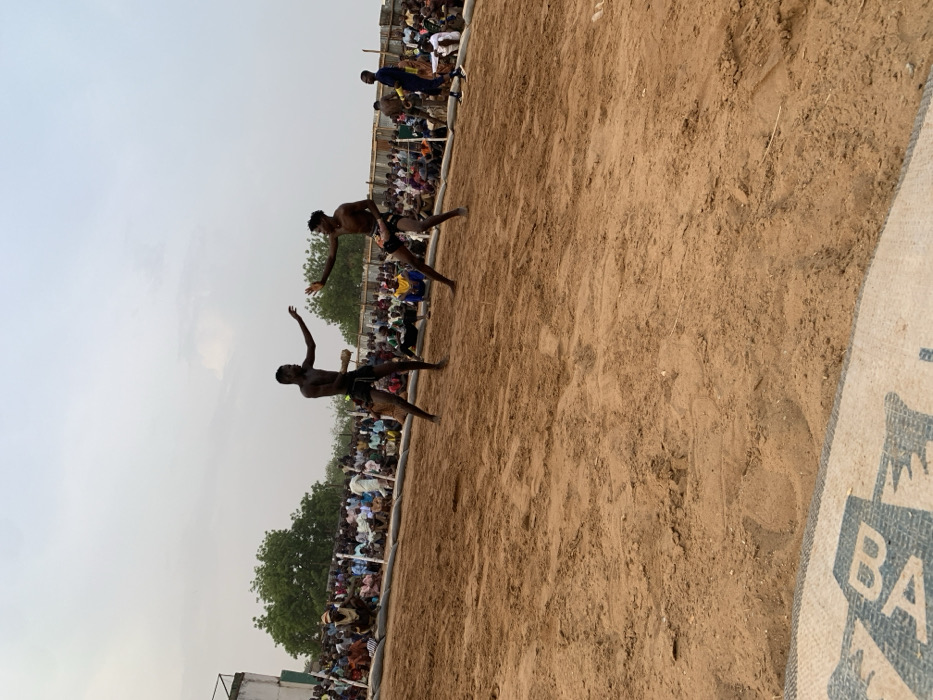 The image shows two men wrestling in a sandy arena, located in the city of Sokoto, Nigeria, at the coordinates 13.049 latitude, 5.238 longitude. The men are shirtless and wearing traditional wrestling attire. One man is holding a stick in his right hand, while the other is striking with his left hand. A large crowd of spectators is seated around the perimeter of the arena. The ground is sandy, and there are some bags in the foreground. The sky is overcast. The men are facing each other, their stances are aggressive. The crowd is watching intently, some clapping and cheering. The overall scene is a traditional wrestling match.