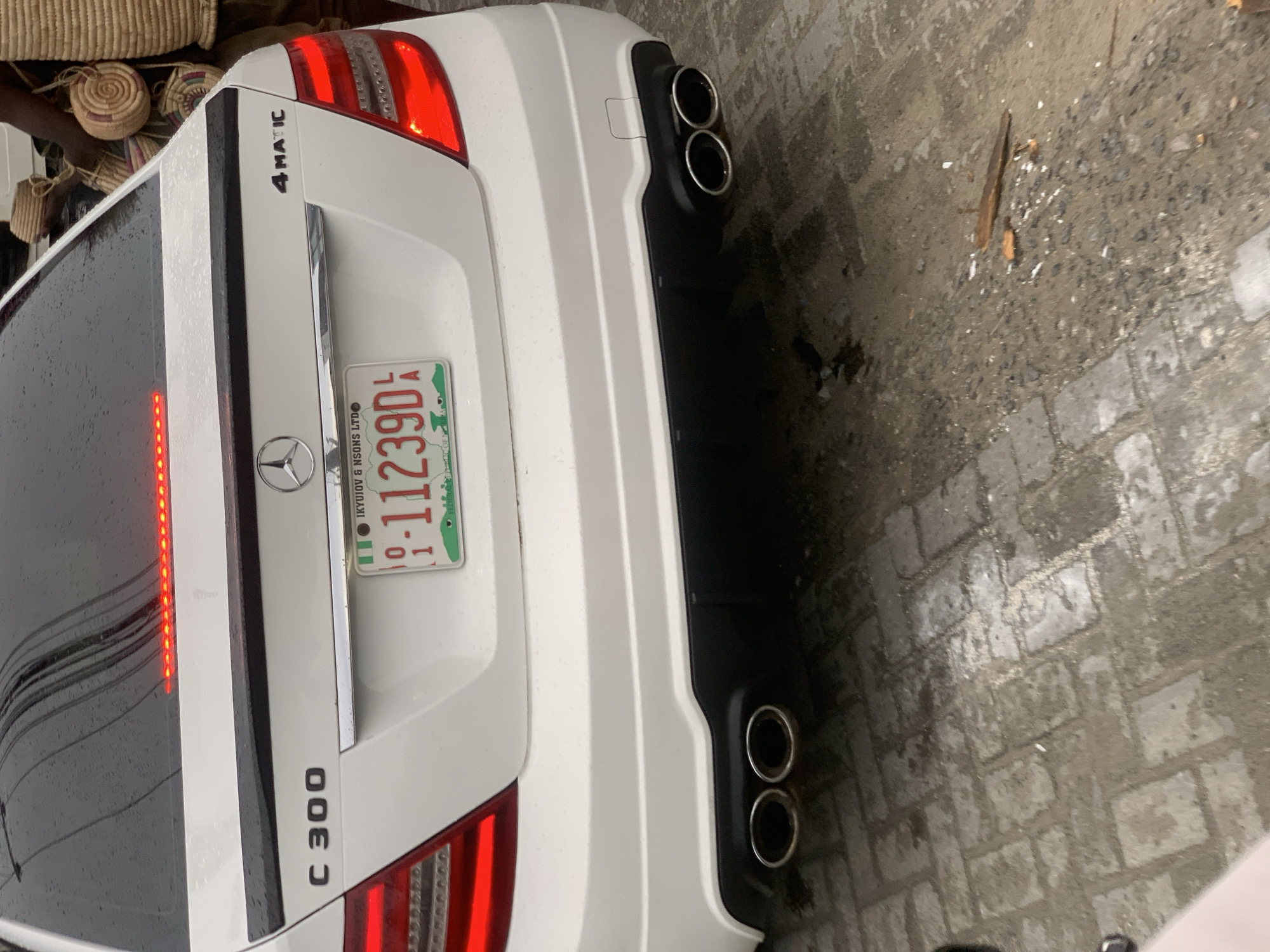 The image shows the rear of a white Mercedes-Benz C300 4MATIC sedan. The car has a Nigerian license plate with the registration number "01-11239D" and the issuing city on the plate reads "LAGOS". The license plate also mentions "IKUYIOV & NSONS LTD". The car has dual exhaust pipes on each side of the rear bumper. The car is parked on a paved surface. Given the license plate mentions LAGOS, the city in which the photo was taken might be Lekki, Nigeria, although this cannot be determined with certainty only from this image.