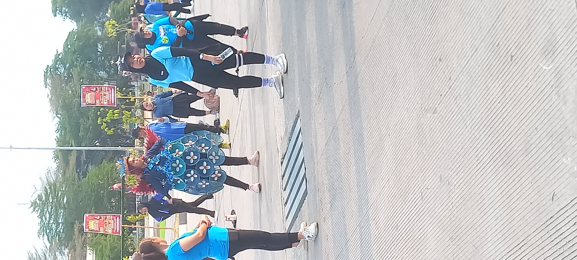 The image shows an outdoor scene, likely in Melayu, Indonesia, where a group of people are gathered. In the center, a person is wearing a distinctive costume that includes a headdress with red feathers and a blue, circular skirt decorated with white flower-like shapes. This individual appears to be the focus of attention, possibly during a performance or event.
Around this central figure, other individuals are dressed in athletic attire, suggesting a recreational activity such as exercise or a community gathering. Several women are visible wearing bright blue tops and dark leggings, some with headscarves. One woman in the foreground on the left is wearing a bright blue t-shirt and black leggings, with white sneakers. Another woman to the right, also in athletic wear and a headscarf, holds a water bottle. In the background, more people are present, some standing and observing.
The environment is paved with a concrete surface that has a textured, ribbed pattern. There is a metal grate or drain cover in the foreground. In the background, trees and banners are visible, indicating a park or public space. The banners have text and graphics, but are not clearly legible. The overall impression is one of a lively community event taking place in an urban outdoor setting.