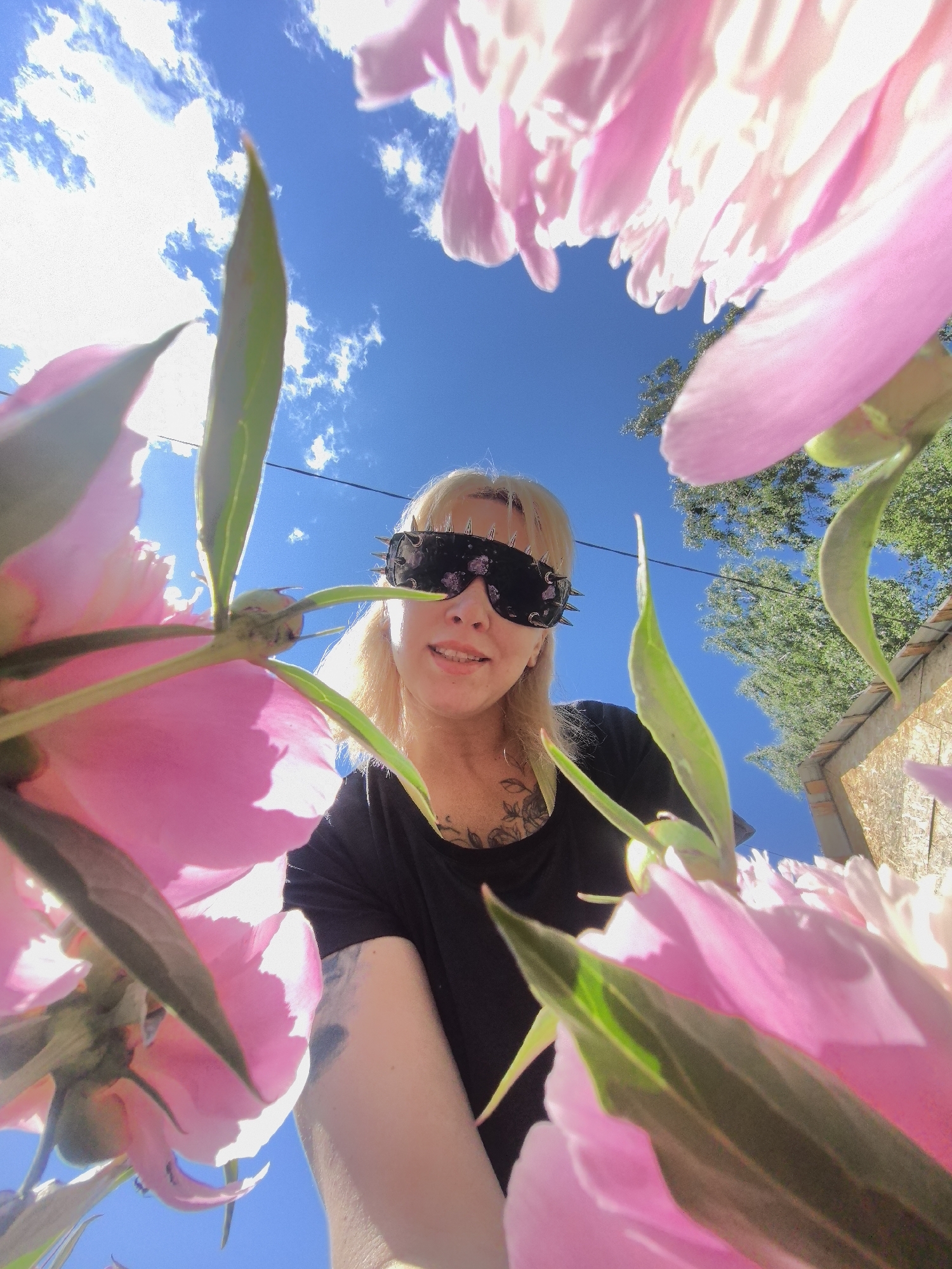 A woman with blonde hair and dark, spiked sunglasses is positioned in the lower half of the frame, looking up at the camera. She is wearing a black t-shirt, and has tattoos on her arm and chest. Large, pink peony flowers fill the foreground and surround her, creating a framing effect. The background consists of a bright blue sky with scattered white clouds and some green foliage, suggesting an outdoor setting. The perspective is from below, looking up through the flowers. The location is specified as Noviy, Russia.