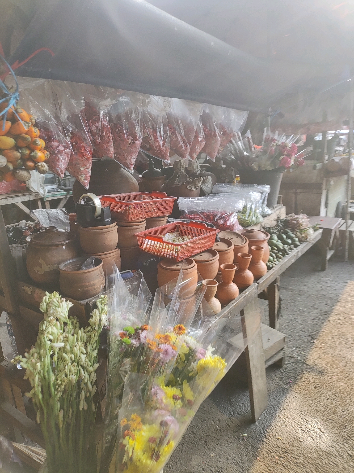 A street vendor's stall in East Jakarta, Indonesia, displays a variety of goods. In the foreground, bouquets of white and multi-colored flowers are wrapped in plastic. Behind them, various clay pots and jars are stacked. The stall is covered by a dark tarp, under which are bags of red petals and clusters of orange betel nuts. The surface of the stall and the ground are visible, with the ground appearing to be a paved or asphalt surface. Sunlight filters through the tarp, creating a bright, diffused light. There are no people clearly visible in the image.