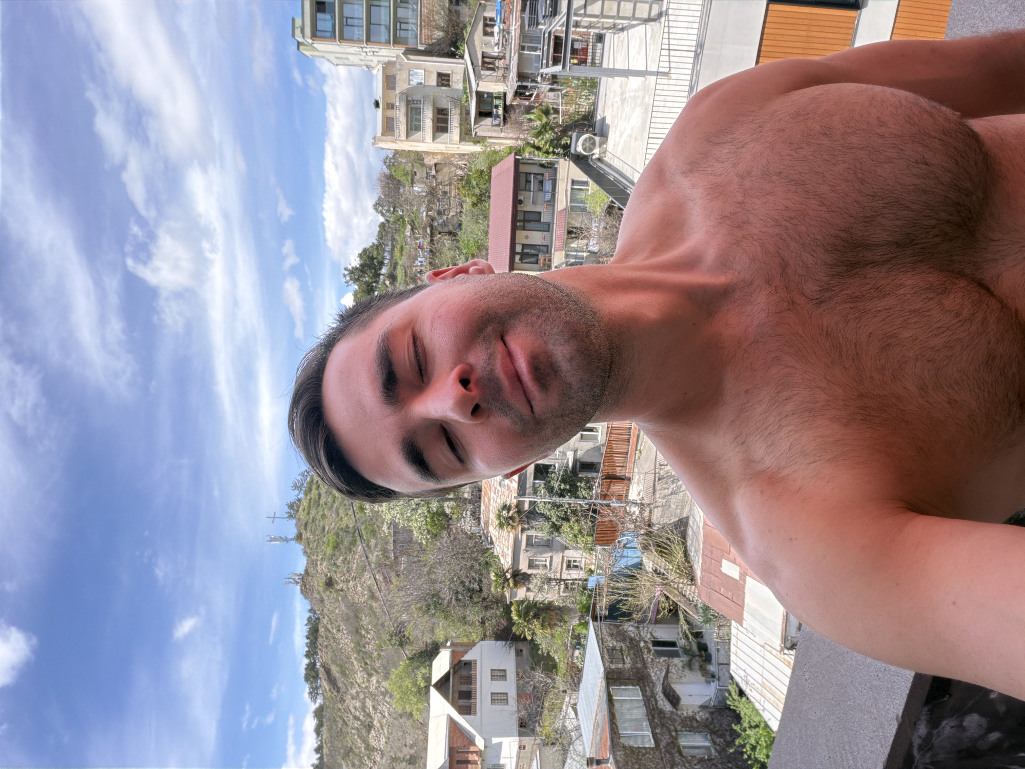 A shirtless man is posing for a selfie. His eyes are closed. Buildings of Tbilisi, Georgia are in the background, as well as a hillside with a cross on the top. The sky is blue with wispy clouds.