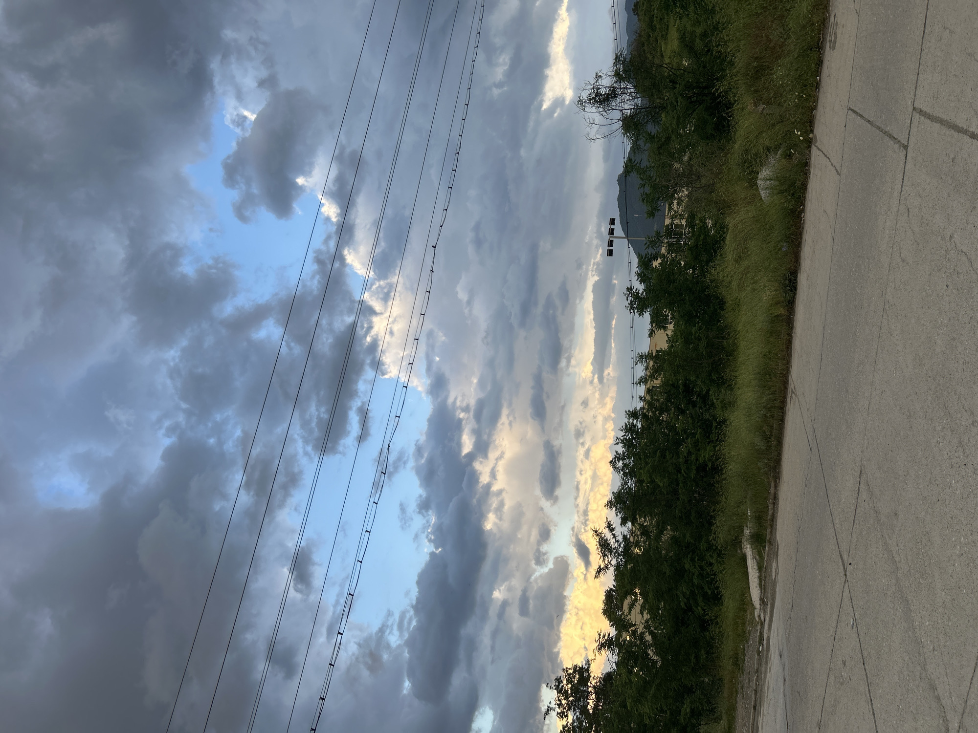 The image depicts an outdoor scene with a view of the sky and a portion of the ground. The sky is filled with a mix of dark, grey clouds and patches of blue sky. Several power lines are visible, stretching across the upper left portion of the image. On the right side, there is a strip of land covered in vegetation, including trees and grass. A paved surface, possibly a road or sidewalk, is visible on the lower right. A few buildings can be seen in the distance, partly obscured by trees. The location appears to be on the outskirts of a city in Unknown, Unknown.