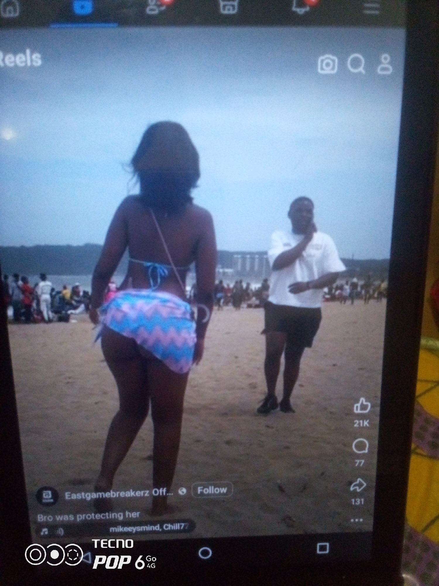 The image displays a TikTok interface on a dark-framed device, with the content showing a beach scene. In the foreground, a woman wearing a blue bikini top and a patterned skirt is viewed from behind, her back towards the camera. To her right, a man in a white t-shirt and dark shorts stands facing the camera, appearing to gesture towards his face. In the background, a crowd of people are visible on a sandy beach with the ocean in the distance, along with some structures that might be buildings or piers. The interface elements indicate a video with a "Reels" header, interaction icons for likes (21K), comments (77), and shares (131), and a caption that reads "Bro was protecting her" with usernames "Eastgamebreakerz Off..." and "mikeeysmind, Chill77". The bottom of the screen shows "TECNO POP 6 Go 4G". The reference to "Howayi, Nigeria" is not present in the visual data of the image.