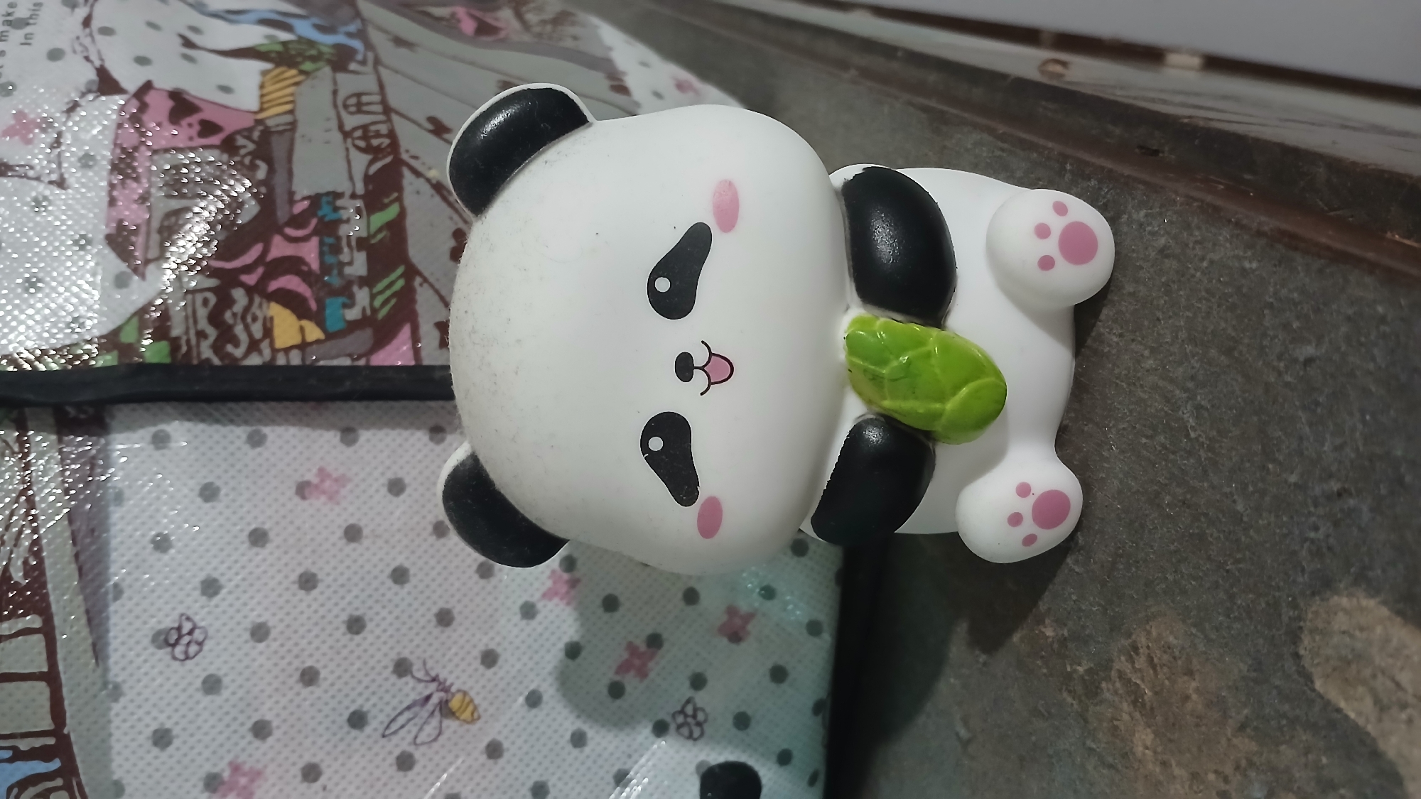 The image shows a close-up of a white and black panda toy sitting on a dark, textured surface. The panda has black ears, black eye patches, pink cheeks, and a small pink mouth. It is holding a green bamboo stalk in its paws. The background is blurred and appears to be a patterned fabric, possibly part of a bag or blanket, with small bee and flower motifs. There is also a portion of a colorful, patterned design visible in the upper right background. The lighting suggests an indoor setting. No specific location in Sampang, Indonesia, can be determined from the image.