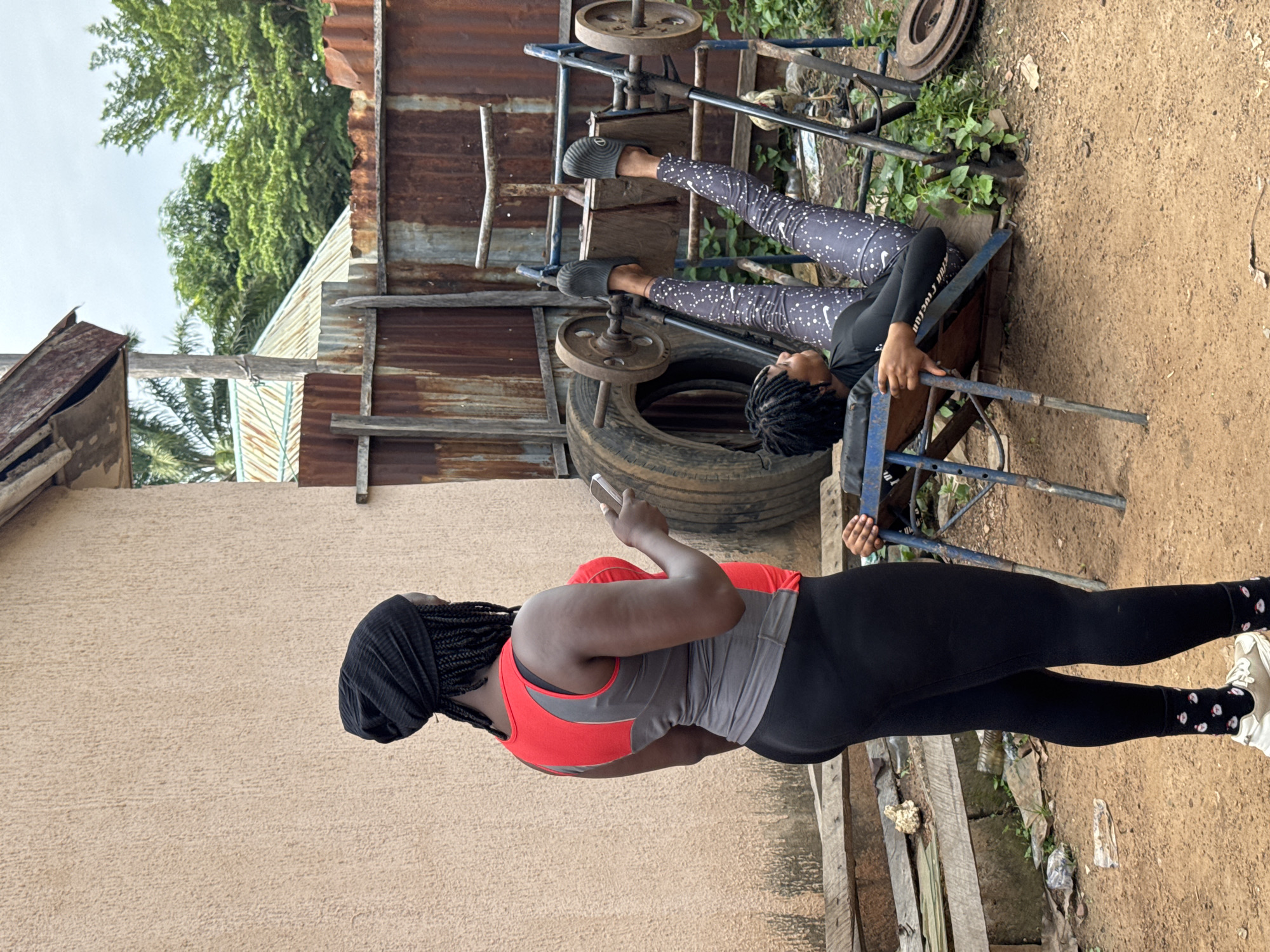 An outdoor scene in Akwanga, Nigeria, features two women engaged in a workout session. In the foreground, a woman stands facing away from the camera, wearing a black beanie, a red and grey athletic top, and black leggings. She holds a mobile phone in her right hand. To her right, a woman lies on a homemade leg press machine, executing a leg press exercise. She wears a black long-sleeved shirt and patterned leggings. The environment is rustic, with corrugated metal fencing, discarded tires, weights, and various other pieces of gym equipment scattered around. The ground is dirt.