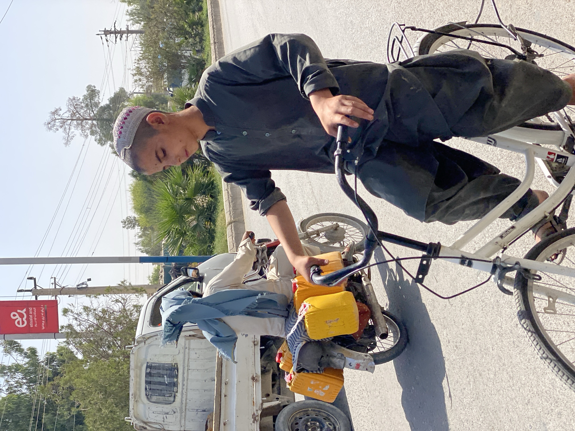 In Kandahar, Afghanistan, on a bright, sunny day, a young boy, likely a teenager, rides a white bicycle on a paved road. He wears a traditional grey tunic and baggy trousers, along with a white and pink embroidered cap. The boy’s gaze is directed downwards. Behind him, to his left, a motorcycle carries a passenger draped in a blue shawl, also wearing a light-colored tunic and trousers. The motorcycle is laden with five large yellow jerrycans, suggesting a transport of liquids. A white truck is parked on the left side of the road, partially visible. The background features trees, utility poles with wires, and a red billboard with the Etisalat logo. The scene captures a moment of daily life and commerce in the city.