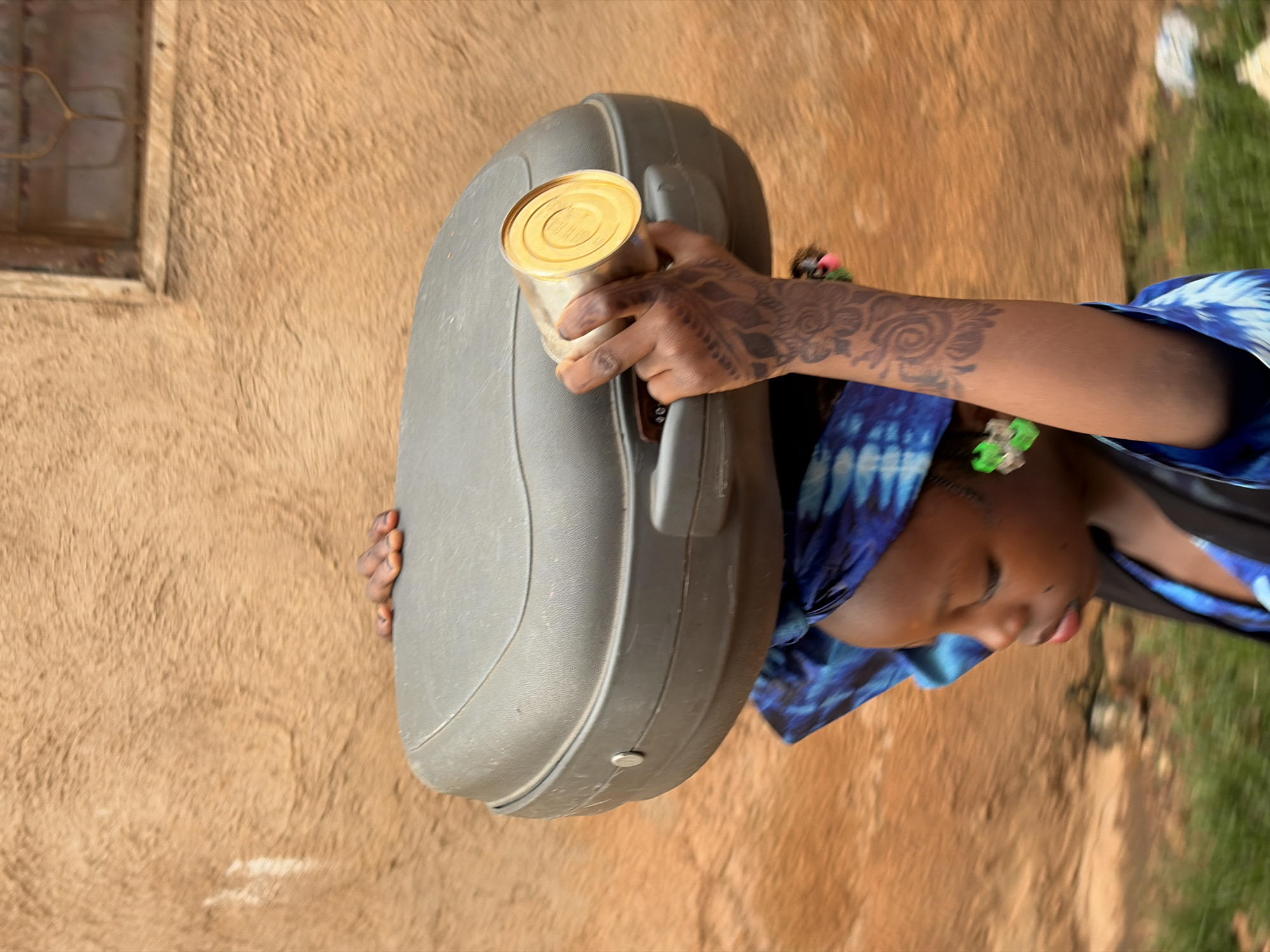 A young person stands outside near a building with a rough, textured beige wall and a window on the left side. The person is holding a gray, hard-shell case with a can resting on it. The person has intricate henna designs on their hand and arm, and wears a blue tie-dye head covering. The location is possibly in Jos, Nigeria.