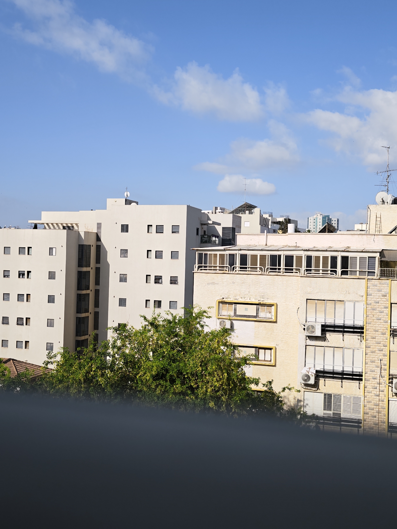 The image captures an urban scene in Petah Tikva, Israel, under a clear blue sky dotted with scattered white clouds. The foreground is dominated by a dark, blurred railing or barrier, suggesting the photo was taken from a vantage point above street level.
The mid-ground features several multi-story apartment buildings. The most prominent building on the left is a modern, light-colored structure with numerous windows, some of which are open, revealing shades or blinds. Its facade is stark and geometric. To its right is another apartment building, slightly taller and with a different architectural style. This building features a prominent enclosed balcony or sunroom with a series of large windows, accented with white railings. The facade of this building appears to be a pale yellow with a textured finish, and some sections display light-colored brickwork. Air conditioning units are visible attached to some of the buildings.
In front of these buildings, a dense canopy of lush green trees provides a vibrant contrast to the urban architecture. The foliage appears healthy and full, with some branches reaching up towards the buildings.
The background reveals more of the city's skyline, including other residential buildings of varying heights and architectural styles. Some of these buildings have antennas or satellite dishes on their roofs. The overall impression is of a densely populated urban environment bathed in natural light. There are no people visible in the frame.