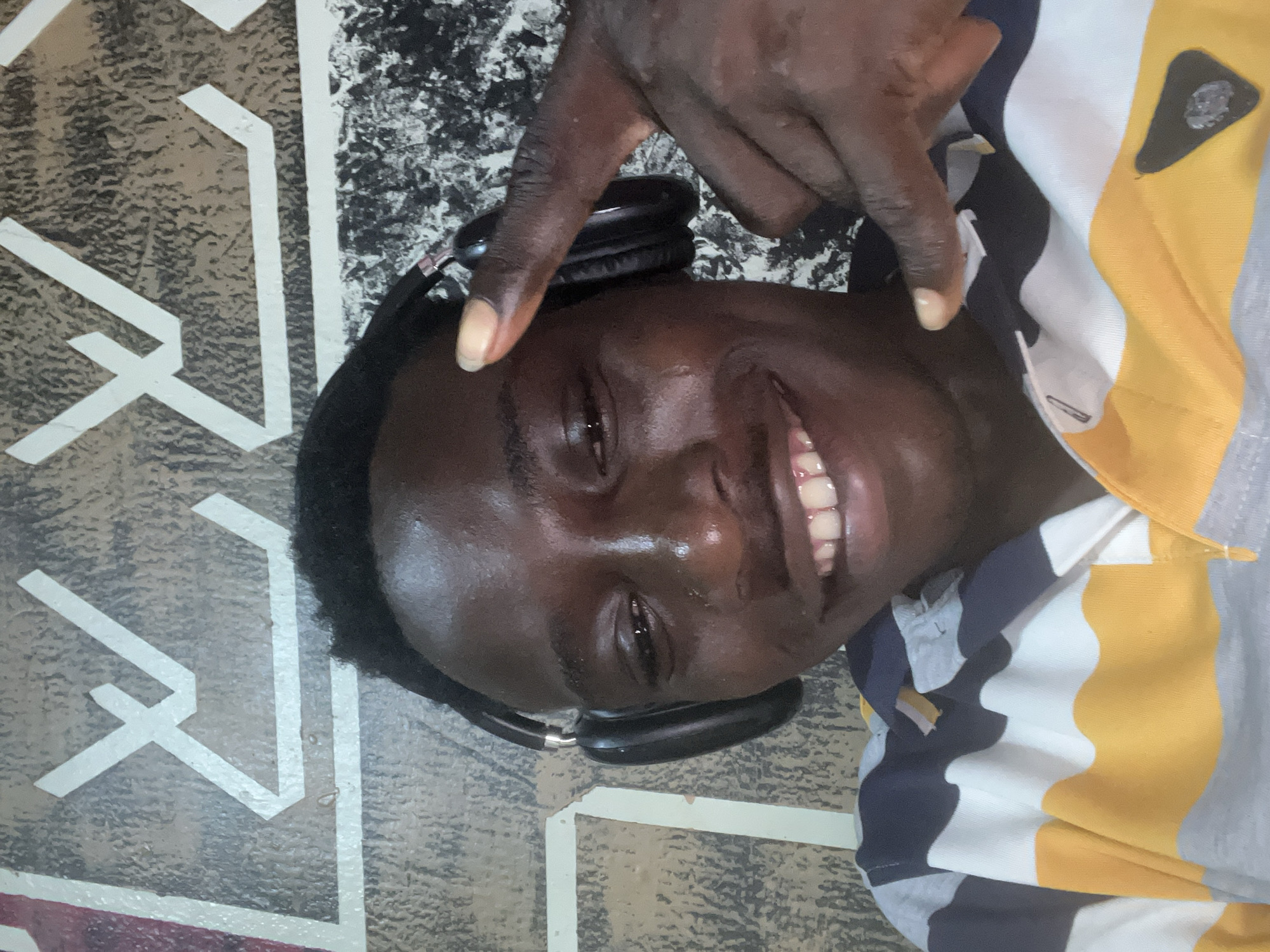 The image captures a young man with dark skin, smiling broadly and looking directly at the camera. He is positioned as if lying down, or the photo is taken from an overhead perspective. He is wearing a multi-striped polo shirt featuring bands of yellow, white, grey, and dark blue or black. A pair of black over-ear headphones rests around his neck.
One of his hands is raised, with the index and middle fingers extended upwards in a 'V' shape, commonly known as a peace sign, and is positioned near his temple. The man's expression is joyful and relaxed, with his smile revealing his upper teeth.
The background consists of a patterned floor or ground surface. To the left of the man, the surface displays a textured greyish-brown color with distinct white geometric lines forming an abstract pattern. Closer to the man's head, the background shifts to a mottled black and white speckled design. The lighting appears soft and even, suggesting an indoor setting or a shaded outdoor area. No specific time of day or weather is discernible, and there is no visible text. The setting for this image is Obosi, Nigeria.