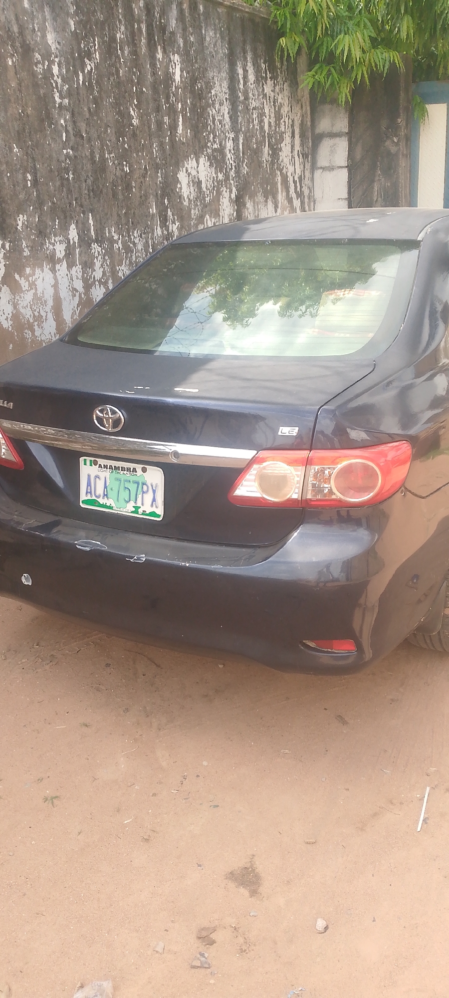 The image shows the rear of a dark-colored sedan parked on a dirt road. The car has a license plate with the text "ACA 757PX" and "ANAMBRA STATE OF THE NATION" visible. The car's taillights are red and white. In the background, there is a weathered concrete wall with patches of white paint and some green foliage at the top. The setting appears to be outdoors, possibly in a residential or semi-rural area, given the dirt ground and the rustic wall. The lighting suggests it is daytime, with some reflections visible on the car's windows. The overall scene is static, with no people or specific activities depicted.