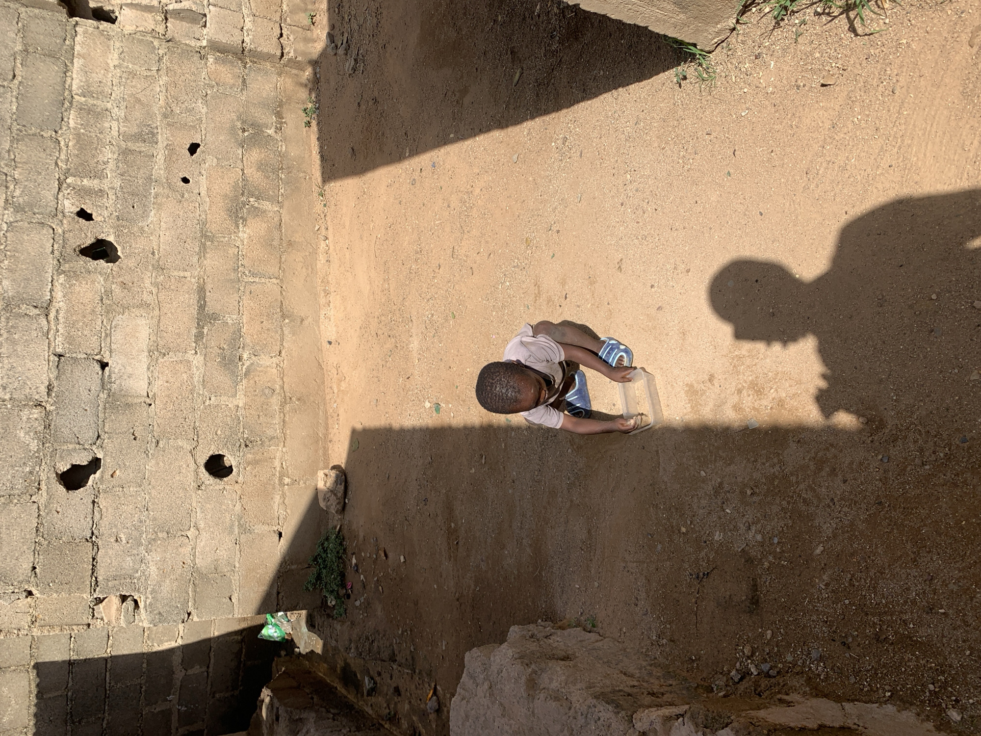 An aerial view shows a young boy holding a translucent plastic container, standing on a sandy ground surface in Rafin Par, Nigeria. The boy's shadow is cast to his right. The sandy ground is bordered by concrete structures, including a wall with holes and damage. Debris and vegetation are scattered near the base of the wall.