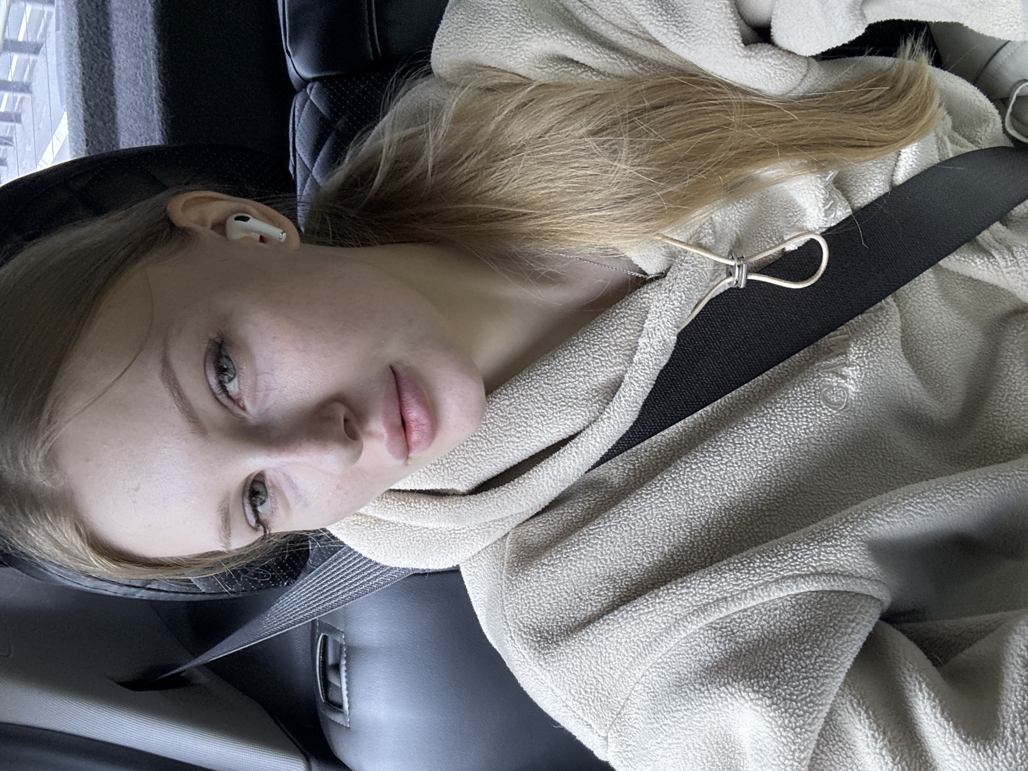 A young woman with blonde hair tied in a ponytail, wearing a light-colored fleece hoodie and a seatbelt, is pictured from the chest up. She has on white wireless earbuds and a necklace. The image appears to be taken inside a car, with the dark interior of the vehicle visible in the background. The scene is indoors, likely in a vehicle. The location is specified as Cheremsha, Russia.