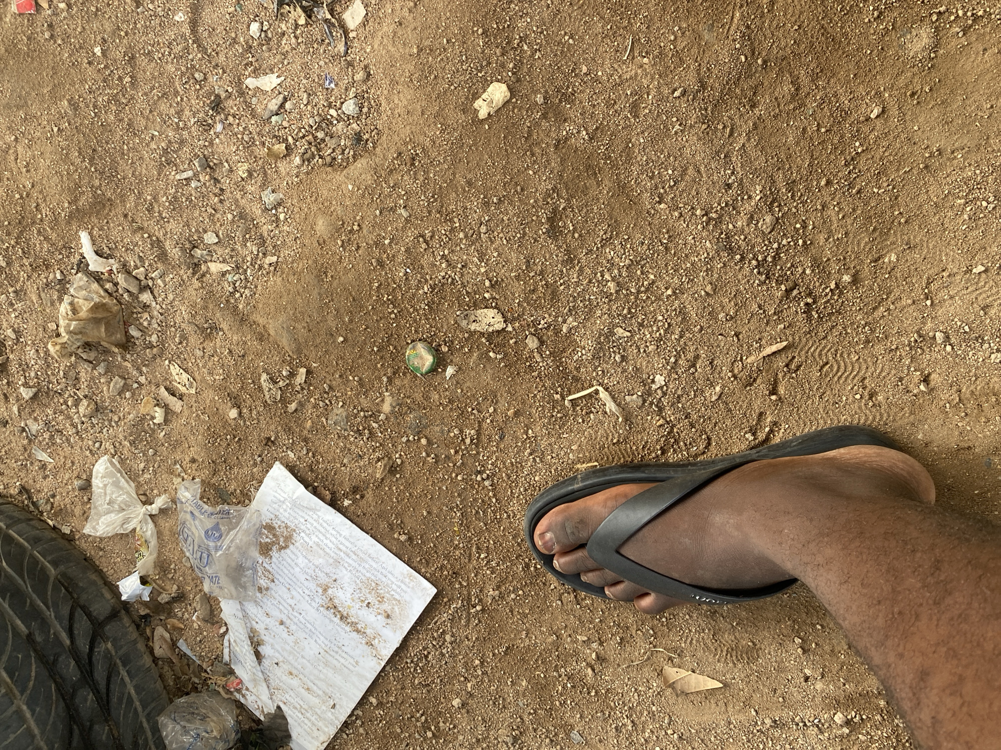 A person's foot, wearing a black flip-flop, is positioned on sandy, dirt ground covered in small pebbles and debris. In the background, a section of a tire is visible, alongside crumpled plastic bags and a white paper document. The scene appears to be an outdoor, possibly urban, setting.