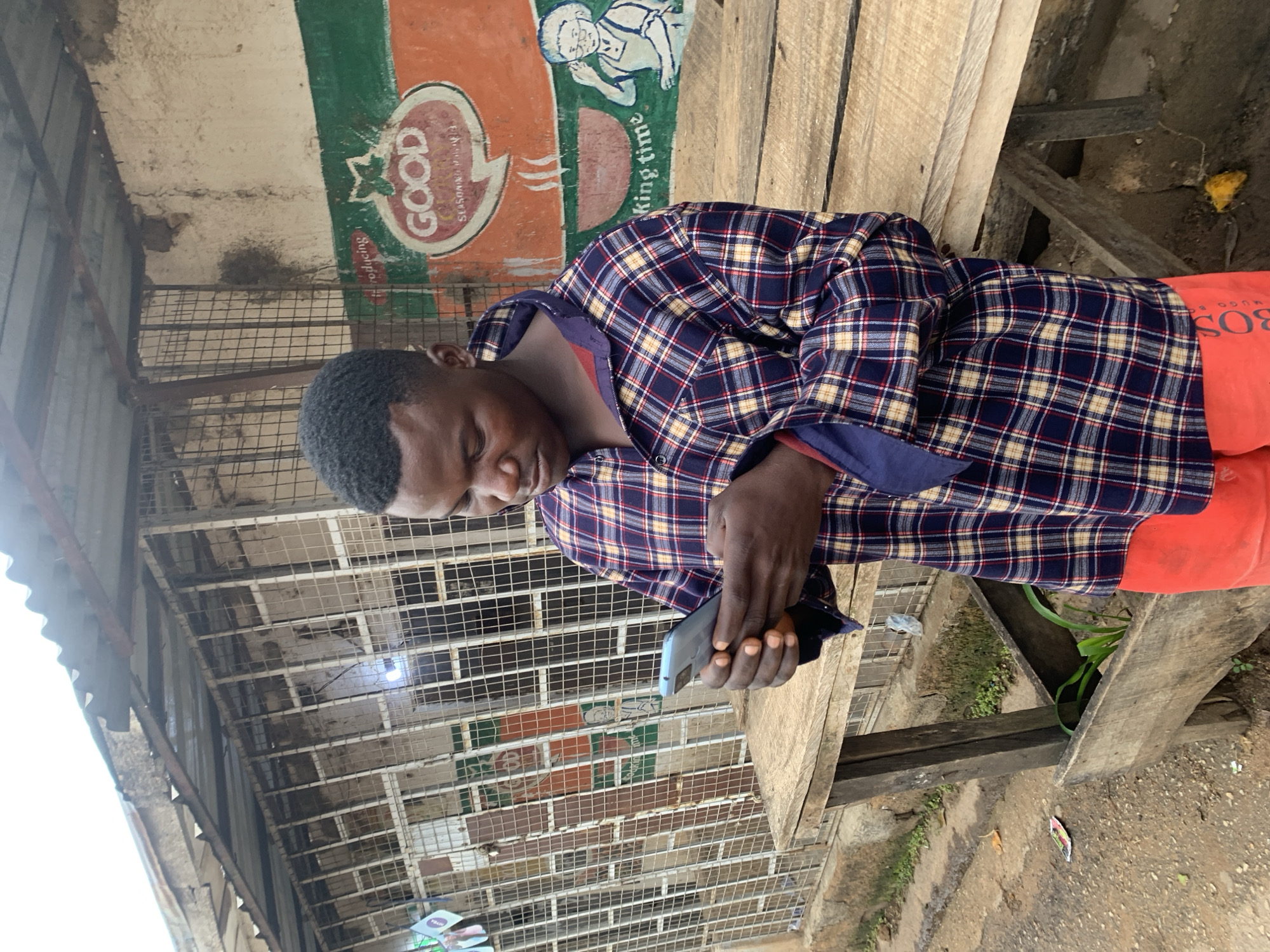 In the city of Jos, Nigeria, a young adult male with dark skin and short dark hair is depicted outdoors in a vertical full-shot, intently focused on a grey-blue smartphone held with both hands. He is wearing a long-sleeved plaid shirt in shades of navy blue, red, and yellow, with the collar slightly open and sleeves pushed up. Below, he wears orange shorts with "BOSS HUGO" visible on the left thigh.
He leans against a rough, unpainted wooden table or stand, its surface composed of multiple planks. Below the table, a trough-like structure contains green plant foliage. The ground beneath is dirt, with scattered small debris including a yellow peel and a wrapper.
In the background, a simple single-story building is visible, featuring a corrugated metal roof. Its front is secured by a white metal grid or gate. To the right of the man, a section of the building's wall displays a painted advertisement: a red tomato illustration above a speech bubble containing the word "GOOD" and "SEASONING" underneath. Below this, "king time" is partially legible, adjacent to a crude drawing of an elderly person. The wall is painted in sections of cream, green, and orange. The scene is illuminated by natural daylight, suggesting an outdoor, possibly urban or semi-urban, setting.