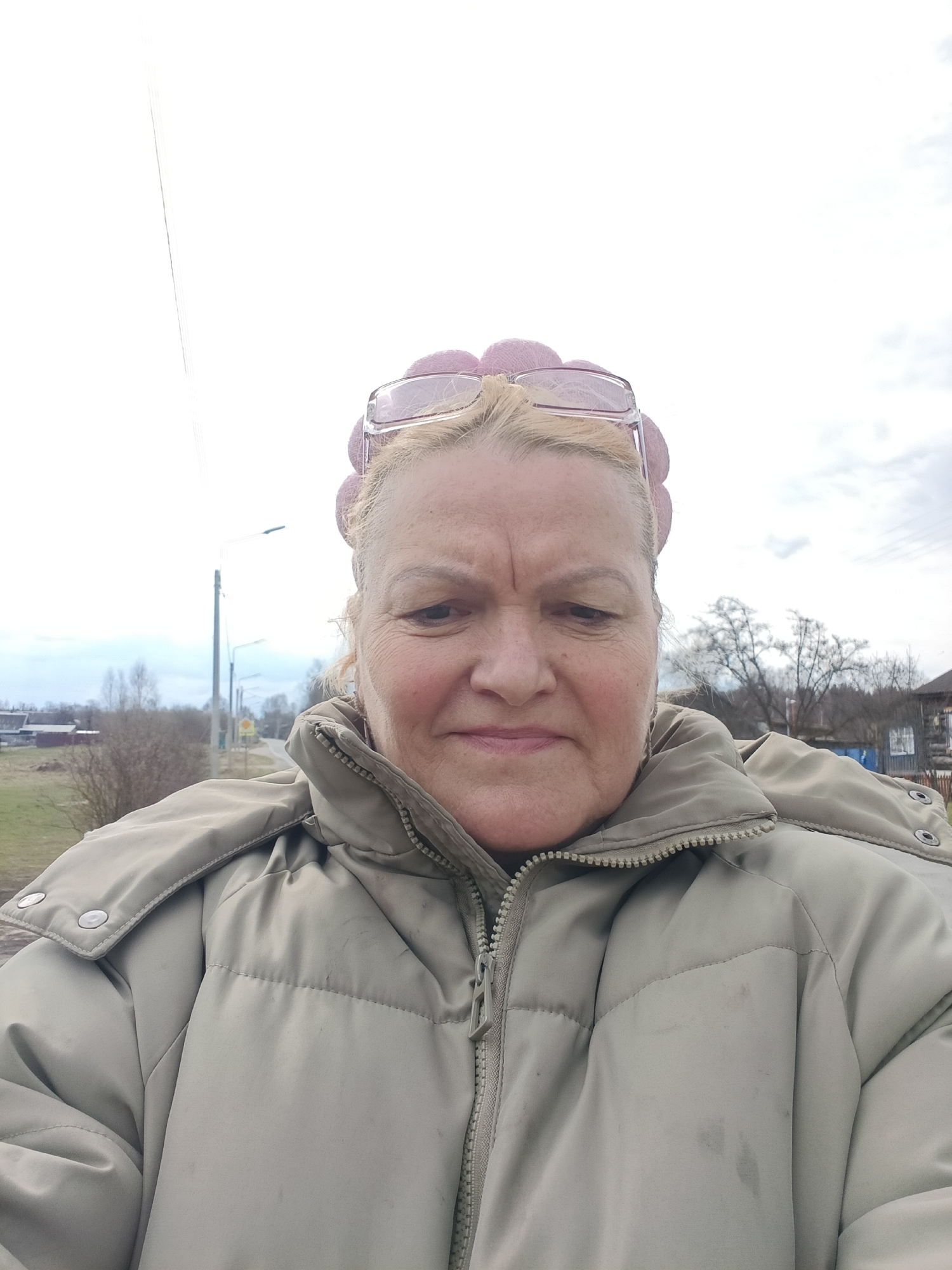 A close-up outdoor selfie depicts a middle-aged to older woman, likely in Krasny Mayak, Russia. She is shown from the chest up, filling most of the frame. Her blonde hair is pulled back, and a pair of pink-framed glasses rests on top of her head. She wears a light greenish-beige (khaki or taupe) puffer jacket with a high zippered collar, and some light stains or smudges are visible on the jacket's front. Her expression is neutral, appearing somewhat thoughtful or weary, with faint lines across her forehead.
The background suggests an overcast daytime setting, possibly in late autumn, winter, or early spring, given the bare trees. To her left, a utility pole stands by what appears to be a road or path extending into the distance, with a field or grassy area beside it. Further in the background on both sides, there are sparse trees and some distant low-rise buildings, indicative of a rural or semi-rural environment. No discernible text is visible within the image.