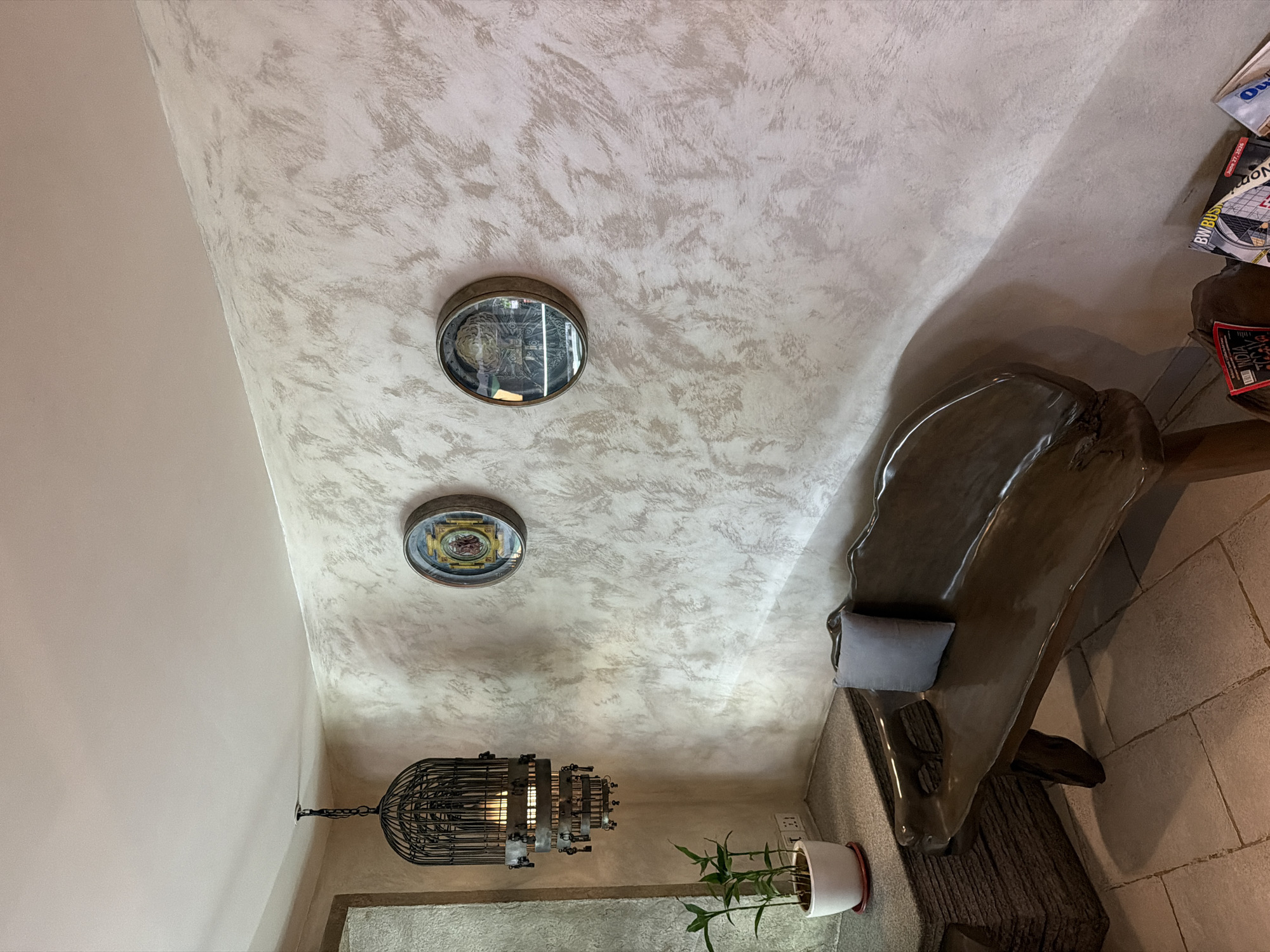 The image depicts an indoor space with a unique ambiance, possibly a waiting area or a lounge. The walls have a textured finish with a light, neutral tone and darker, irregular streaks, giving them a distressed or aged appearance. On the wall, two circular decorative plates with intricate designs are mounted. To the left, a distinctive birdcage-shaped chandelier hangs from the ceiling, illuminated by a warm light. Below the chandelier, on a textured ledge, sits a white pot containing a small green plant with slender leaves. Adjacent to this is a dark, irregularly shaped wooden bench with a smooth, polished finish. A light gray cushion rests on the bench. The floor is tiled with large, light-colored square tiles. In the lower right corner, several magazines are stacked, some with visible text and images. The overall lighting is soft and diffused. The scene is devoid of any people. The presence of magazines suggests a public or commercial space. Given the presence of numerous upscale cafes, boutiques, and artistic installations in areas like Bandra and Colaba, it is plausible this scene is located in a well-appointed establishment within Mumbai, India, designed to offer a visually appealing and tranquil environment for patrons.