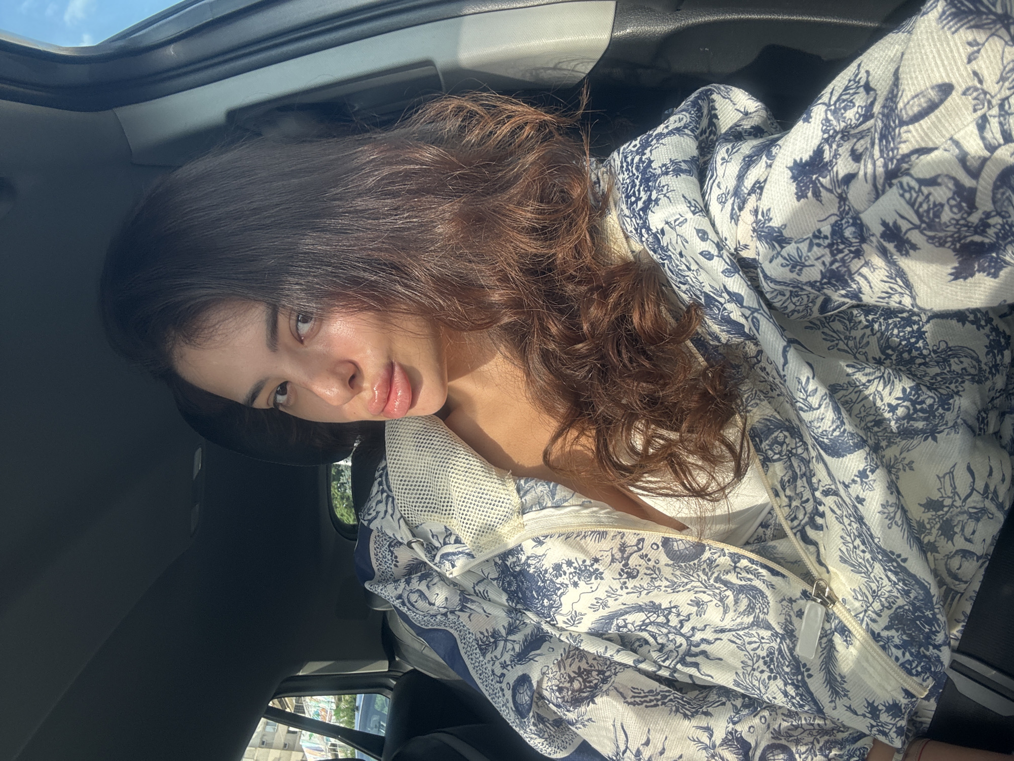 A young woman is sitting in the passenger seat of a car. She has long, wavy brown hair and is wearing a white top and a blue and white patterned jacket. The jacket has a toile de Jouy print. Her lips are slightly parted and her eyes are looking towards the camera. The sun is shining through the car window, casting a warm glow on her face. Outside the car, a cityscape with buildings and trees is visible. The interior of the car shows a dark gray headliner and door panel.