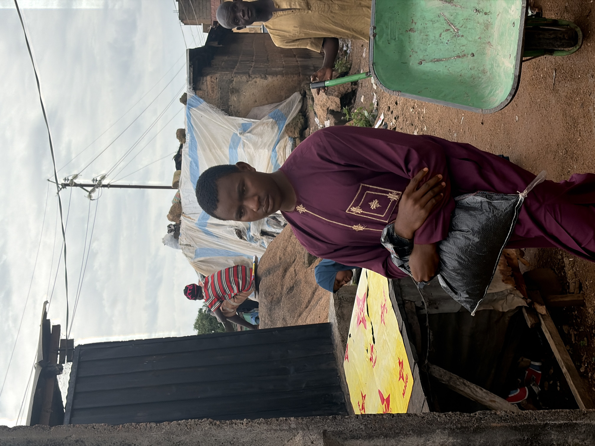 In Jos, Nigeria, a young man in a maroon tunic and pants stands in the foreground, holding a black plastic bag. To his right, another man in a tan, button-down shirt stands near a green wheelbarrow. In the background, a person in a red and white striped shirt and a headscarf sits on a pile of rocks behind a black metal gate. A woman wearing a blue headscarf is visible near a yellow tarp. The environment appears to be an outdoor market or street with rough terrain and makeshift structures covered by tarps. The sky is overcast.