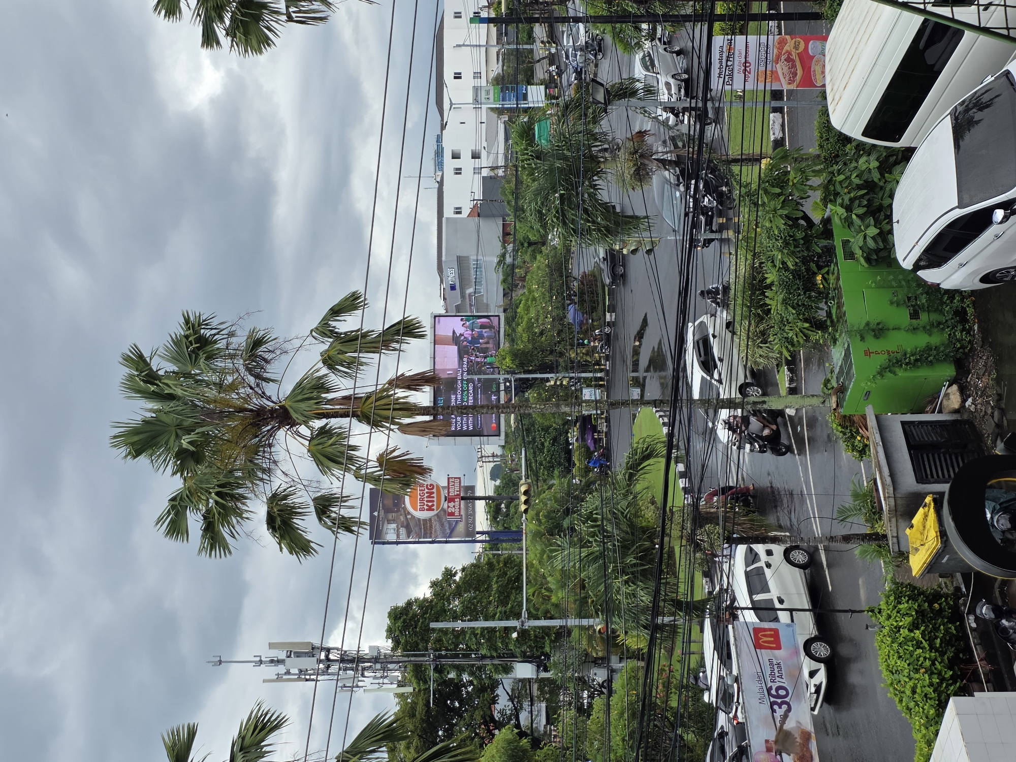 The image depicts a street scene in Kuta, Indonesia, viewed from an elevated perspective. Overcast skies provide a muted backdrop. Lush tropical vegetation, including palm trees, lines the road, interspersed with utility poles and electrical wires that crisscross the view. Traffic, consisting of cars and motorbikes, moves along the wet asphalt of the road. Billboards advertising Burger King, McDonald's, and a fitness center (FINESS) are visible along the roadside. The Burger King billboard includes the text "24 HOURS DRIVE THRU." A large billboard advertising "RIDE ADINE" offers "40% OFF ON GRAB WITH TERCARD." Buildings of varying heights and architectural styles line the opposite side of the street.