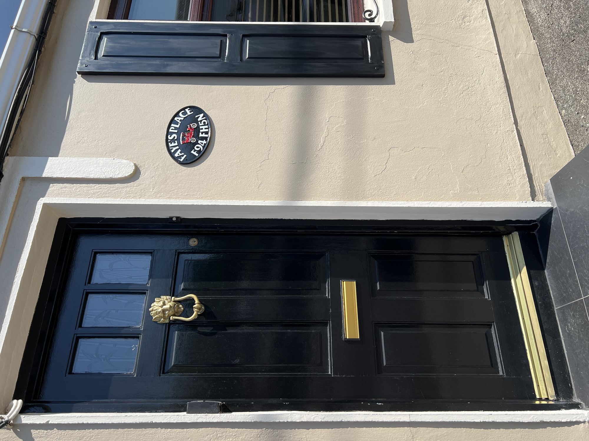 This image depicts the exterior of a building in Donegal, Ireland. The main subject is a black, paneled front door with a gold lion-head door knocker and a gold mail slot. Above the door, there are three small, frosted glass panes with a bamboo pattern. To the right of the door, on the pale beige stucco wall, is an oval black sign with white lettering that reads "FAYE'S PLACE F94 FH5N" and features a red vintage car graphic. Adjacent to the sign is a dark blue louvered shutter covering a window. The building has a white decorative trim around the door frame and a white downpipe visible at the top. The ground in front of the door is paved with dark tiles, and the surrounding wall shows some minor cracks in the stucco. The scene is illuminated by bright sunlight, casting distinct shadows.
