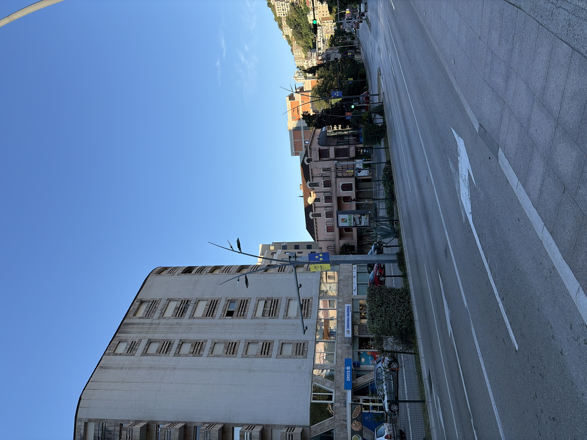 This is a landscape, eye-level, medium shot of a street in Budva, Montenegro, under a clear blue sky. A tall, pale, multi-story building with rows of rectangular windows and balconies curves on the left side of the image. The building's lower level features large reflective windows, revealing the sky and surrounding buildings. A street runs diagonally from the bottom left to the upper right, with white lane markings and an arrow indicating a right turn. Several cars are visible on the street, and a pedestrian walkway with grey tiles borders the road on the right. Across the street, there are more buildings of varying heights and architectural styles, some with red-tiled roofs. Lush green trees and vegetation are interspersed between the buildings and along the hillside in the background. A traffic light with a green signal is visible in the distance. The overall impression is of a sunny day in an urban environment.