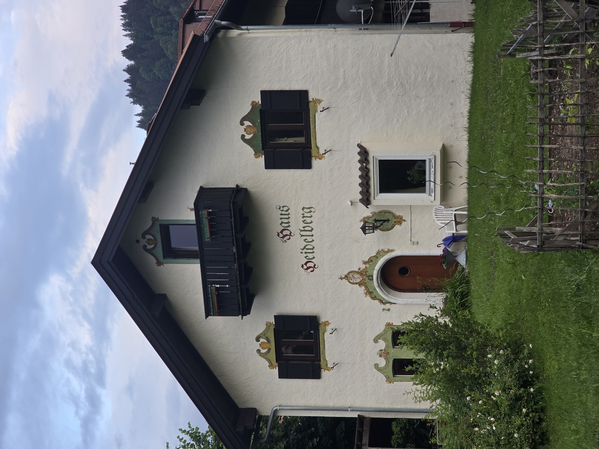 The image shows a house with the name "Haus Heidelberg" written in ornate lettering on its facade, located in Garmisch-Partenkirchen, Germany. The house is light-colored with dark trim and features several decorative elements, including colorful painted embellishments around the windows and the arched wooden door. There are black shutters on some of the windows, which are open. A small black balcony with decorative railings is present above the central entrance. The house is surrounded by lush green grass, and a dense line of trees forms the background, suggesting a semi-rural or mountainous setting. In front of the house, there is a bush with white flowers and a low wooden fence. A white plastic chair and a blue umbrella are placed near the entrance, and a clothes drying rack is visible on the right side of the building. The sky above is cloudy.