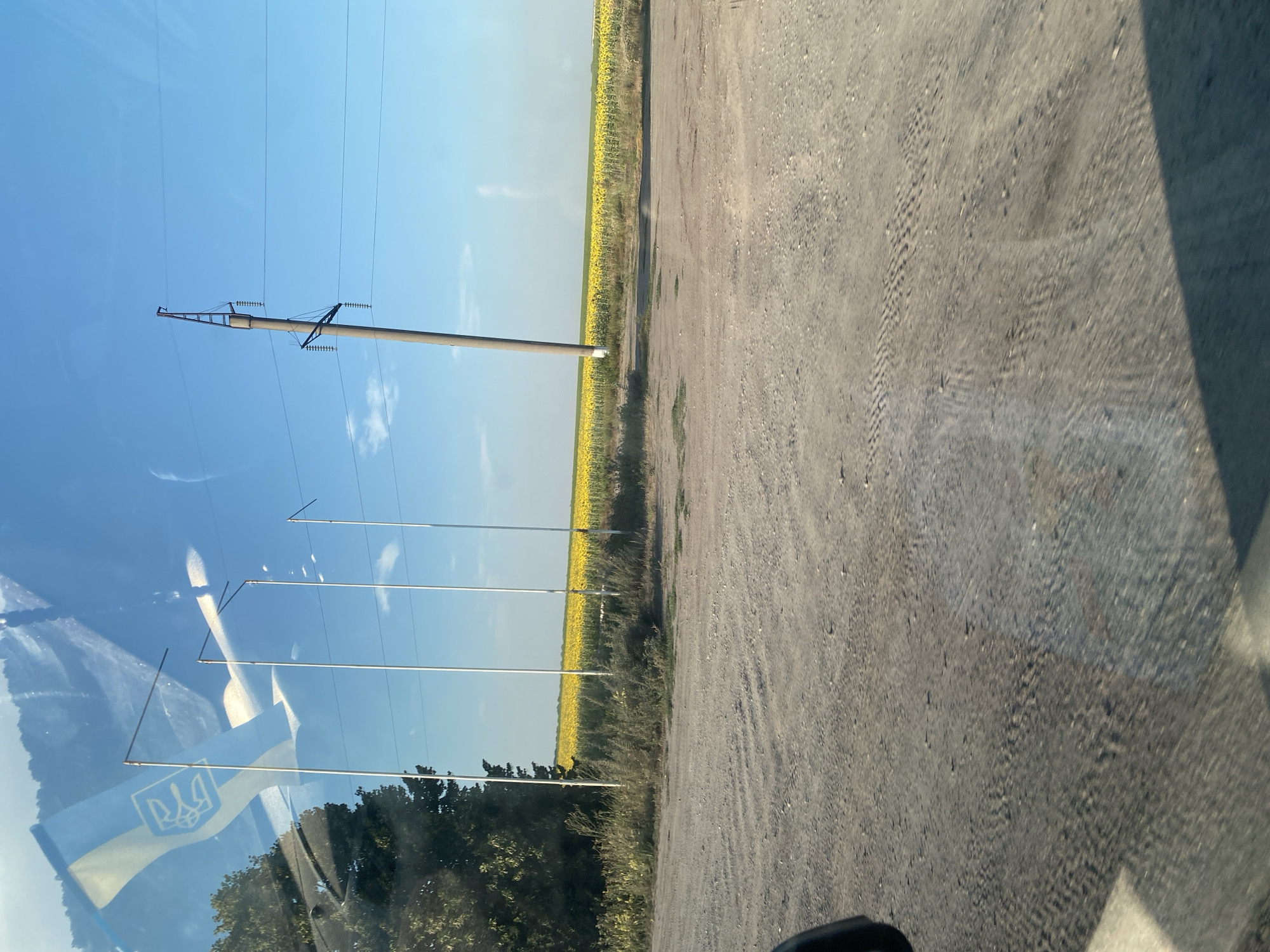 The image depicts a field of sunflowers under a clear blue sky, with a power line pole in the foreground and a dirt road. The reflection of a Ukrainian flag with a trident emblem is visible on a surface, likely a window. Several tall poles, possibly flagpoles, are also present, and a sliver of a tree line is on the left. The overall scene suggests a rural setting in Ukraine, possibly near the town of Maiske.